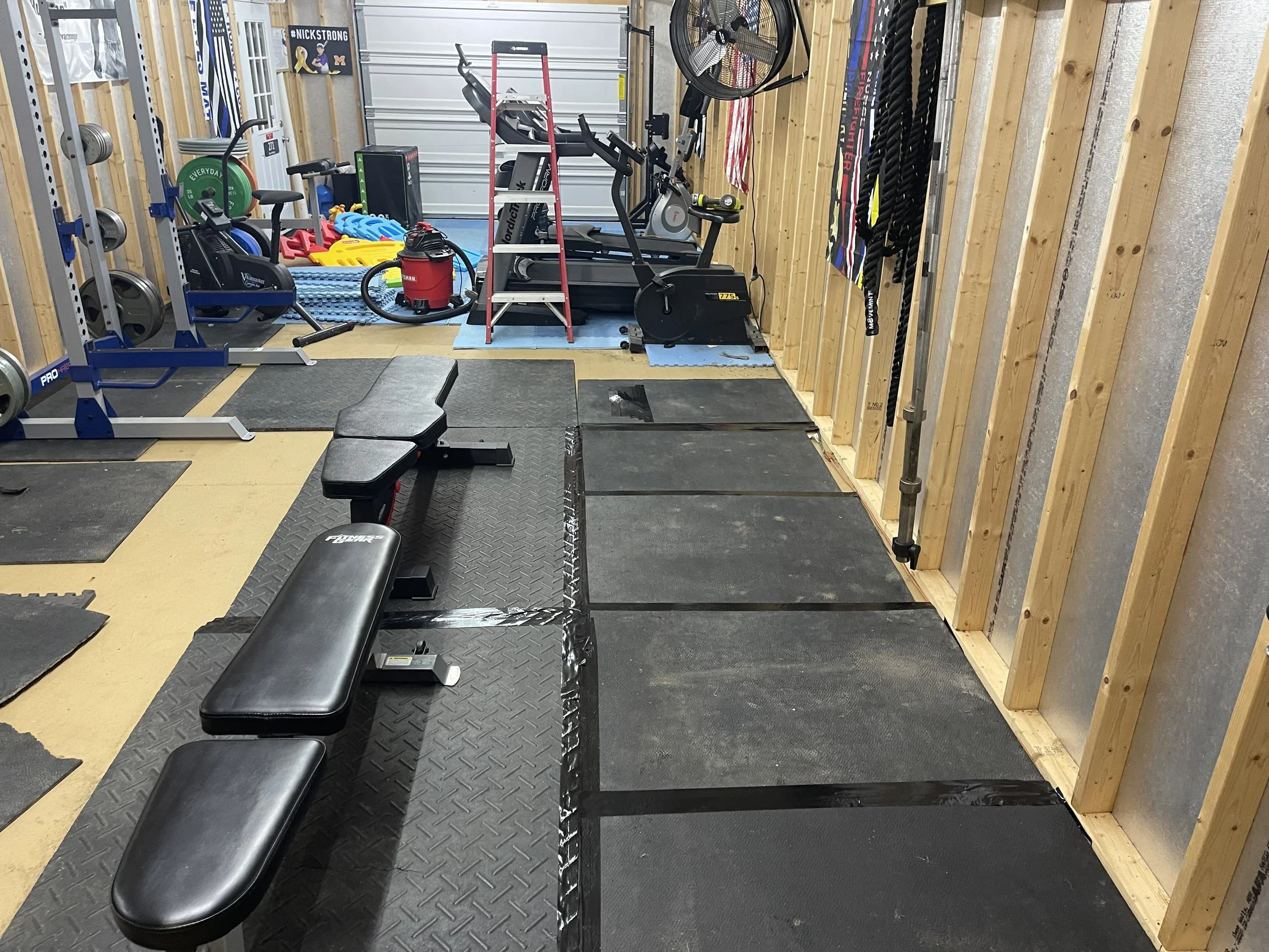 A well-organized home gym with weightlifting equipment, a treadmill, a rowing machine, resistance bands, and various gym accessories in a room with unfinished wood walls.