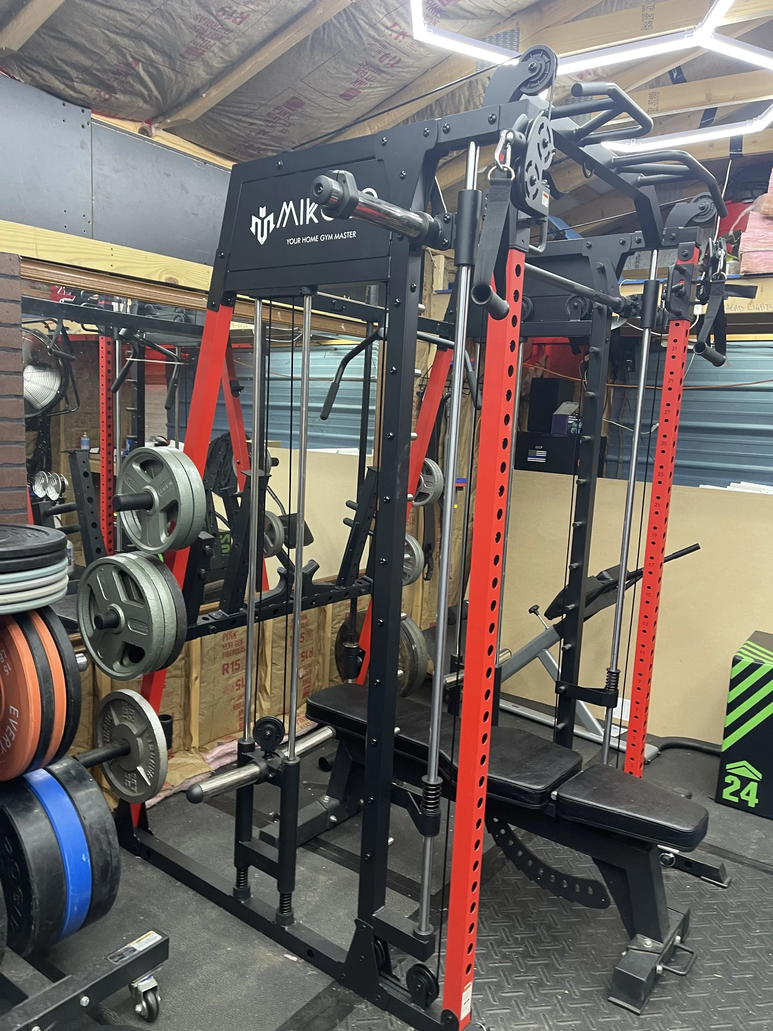 A multi-functional home gym apparatus with weight plates, a bench, and various bars and pull-up bars, in a garage setting with insulation and a brick wall.