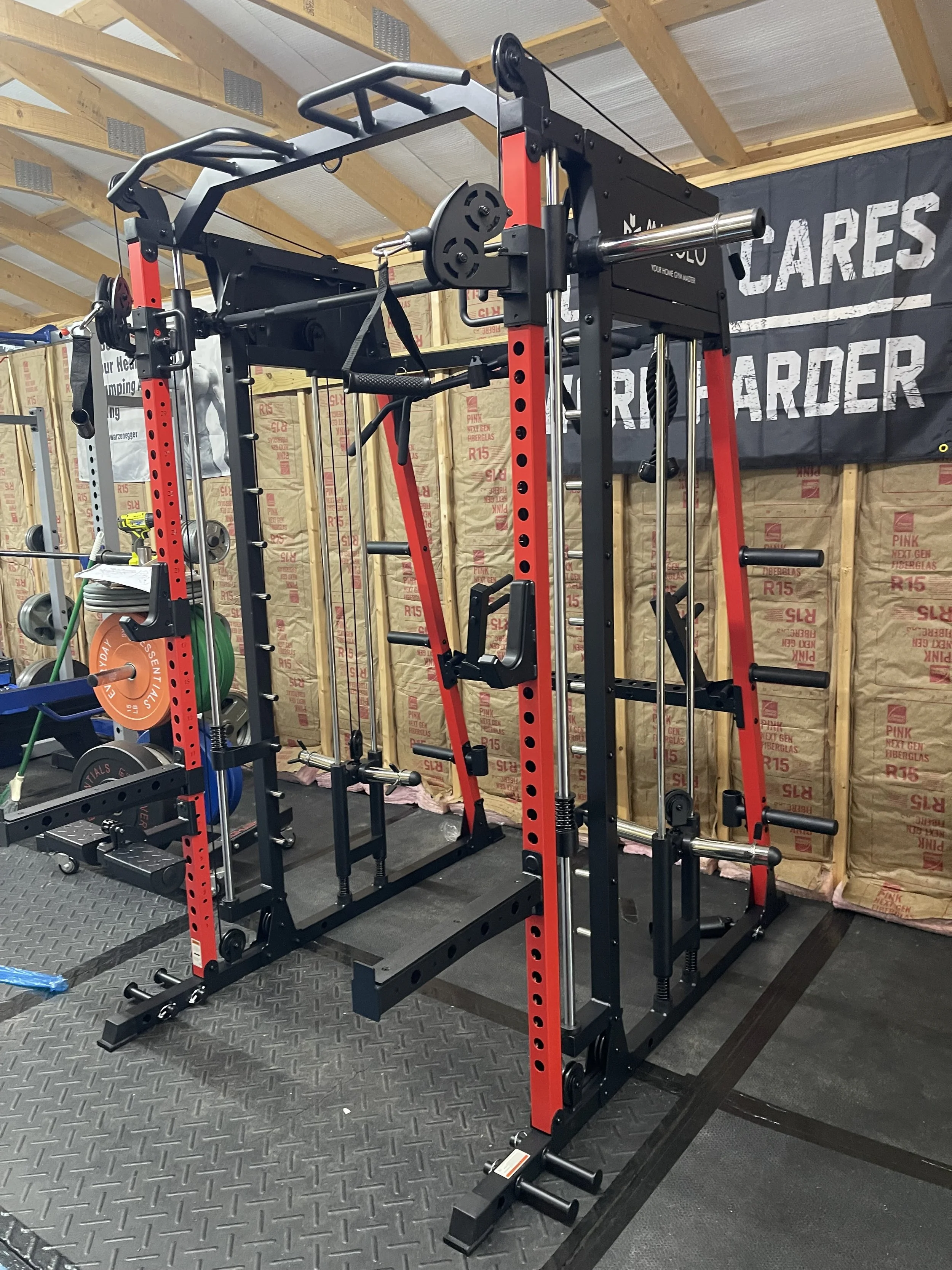 A squat rack and pull-up bar in a gym with insulation on the walls and a banner that says, 'WE CARE, BE STRONGER'.