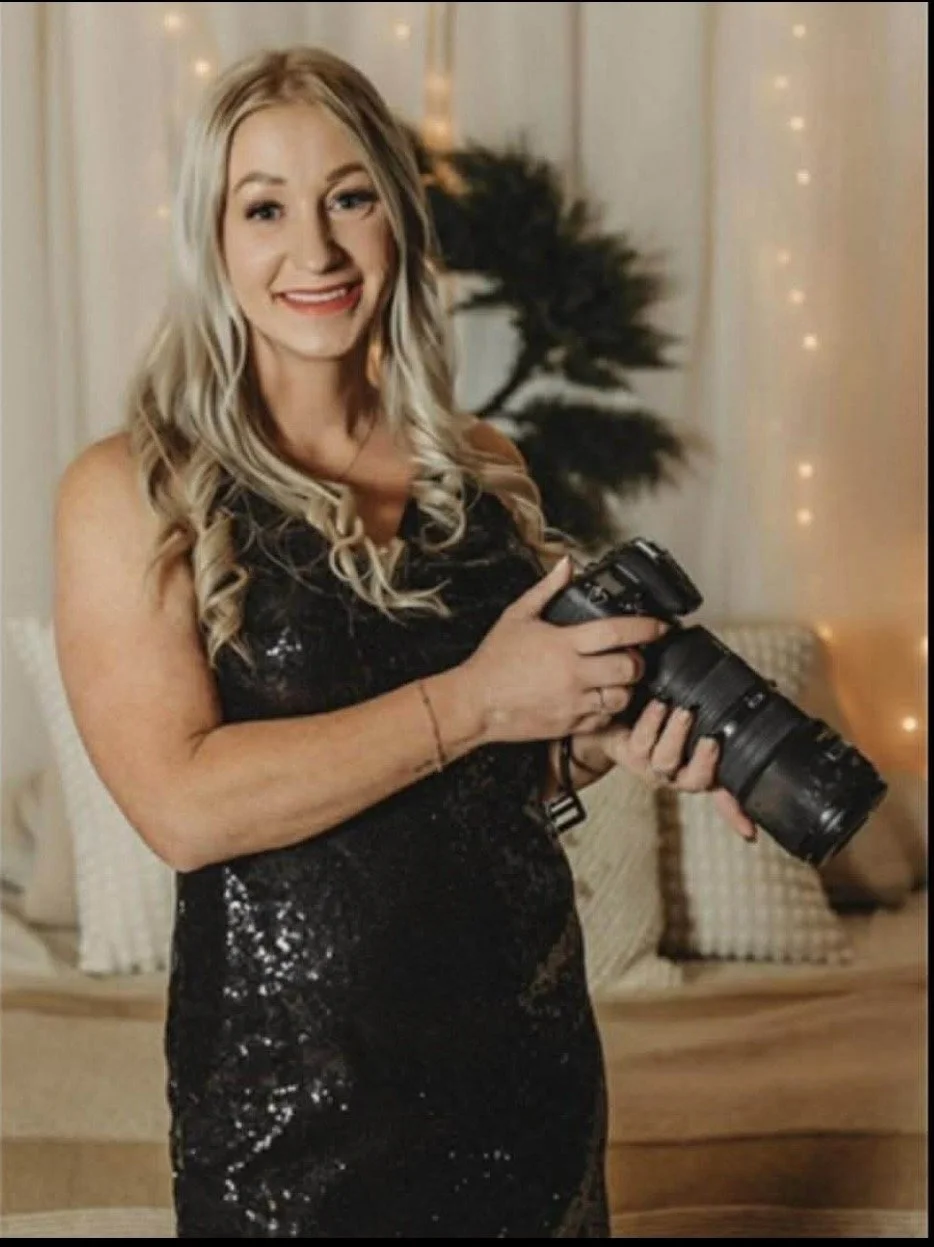 A woman with long blonde hair in a black sequined dress holds a camera and smiles at the camera. The background has a blurred indoor setting with warm lighting and decorative string lights.
