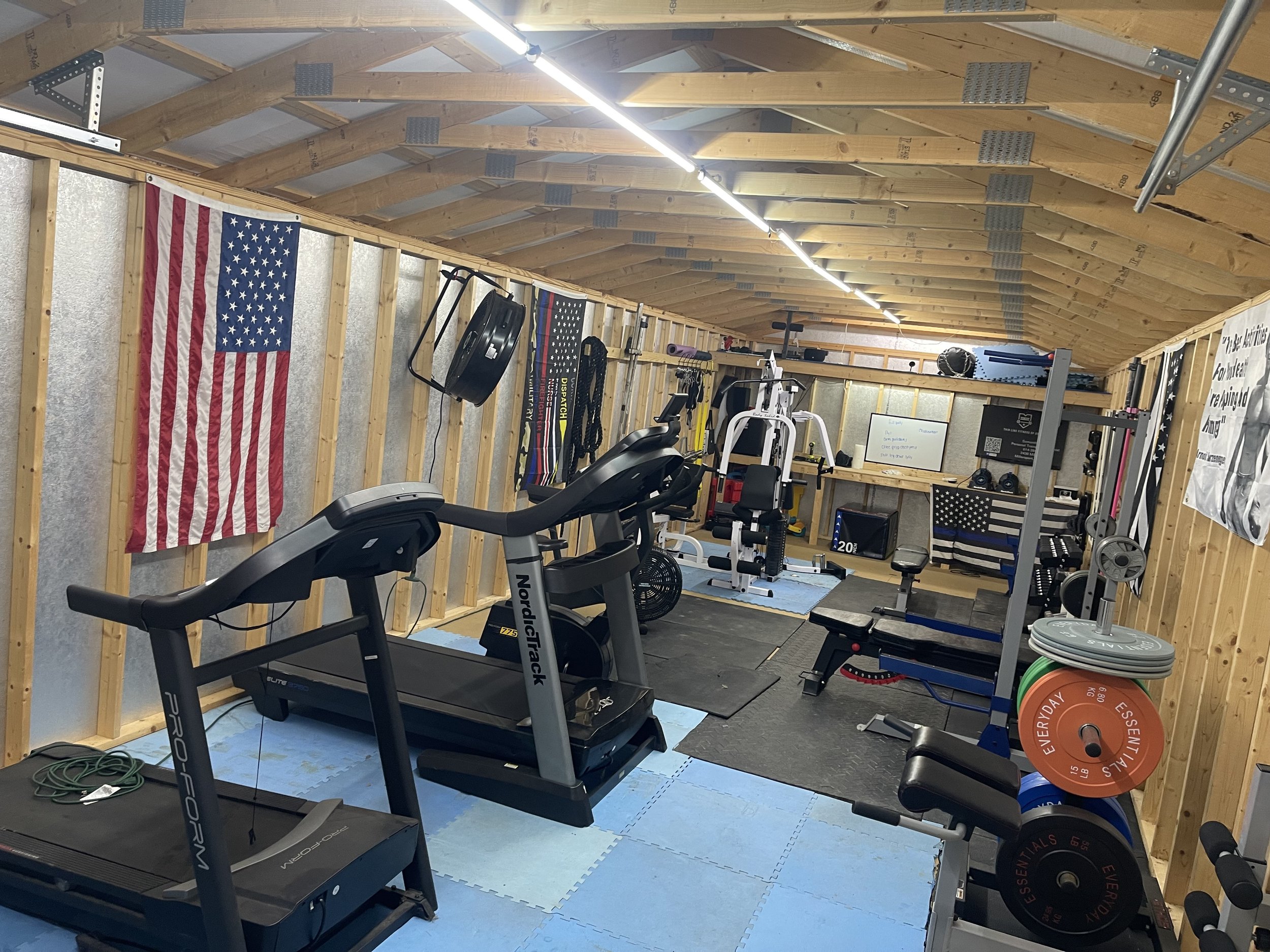 Home gym with treadmill, weightlifting equipment, American flags, and various workout accessories in a wooden-walled room.