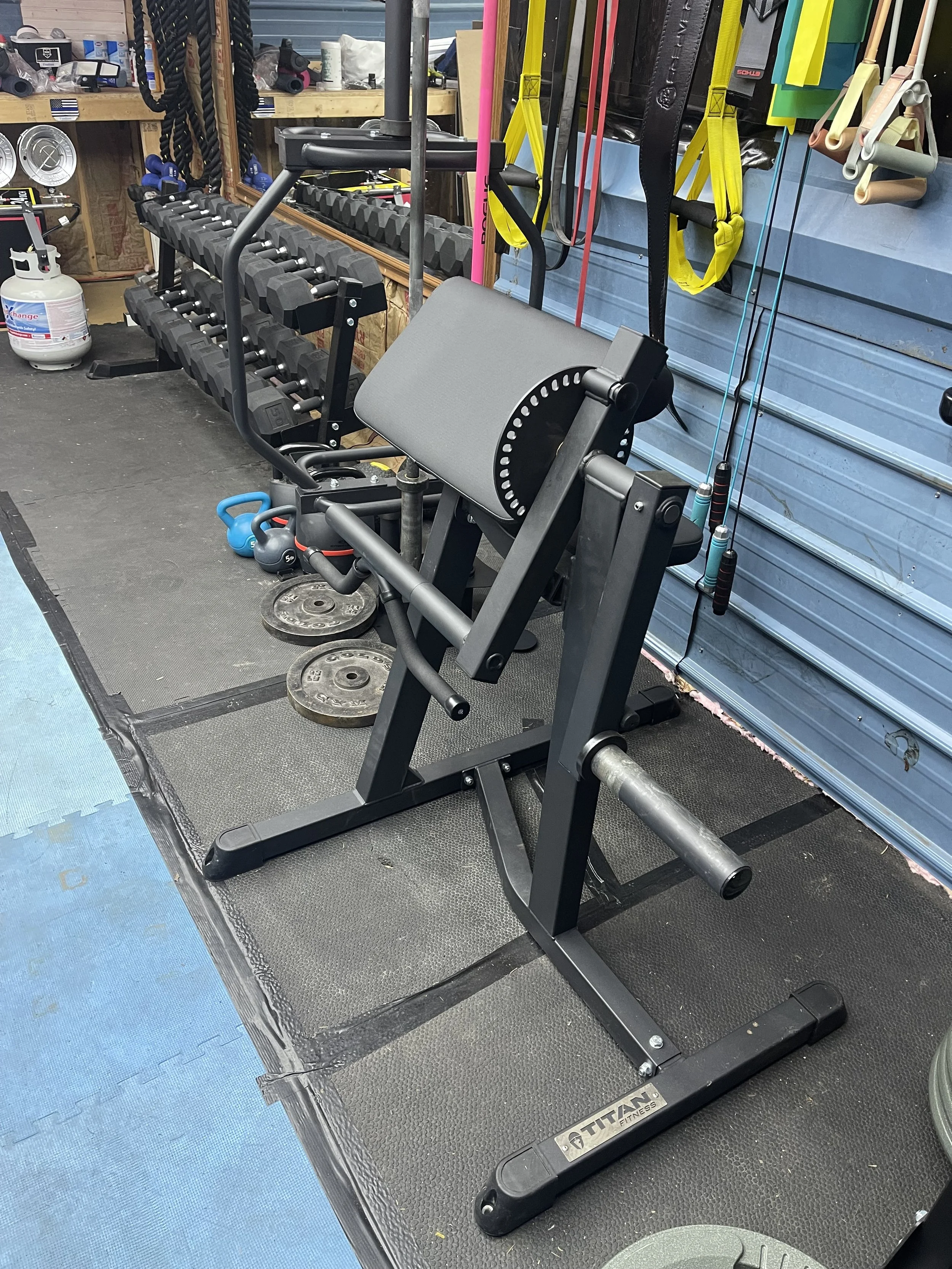 Fitness equipment including a hip abduction machine in the foreground and a row of dumbbells on a rack behind it, with various workout straps hanging on the wall and a propane tank on the left side in a garage gym setting.