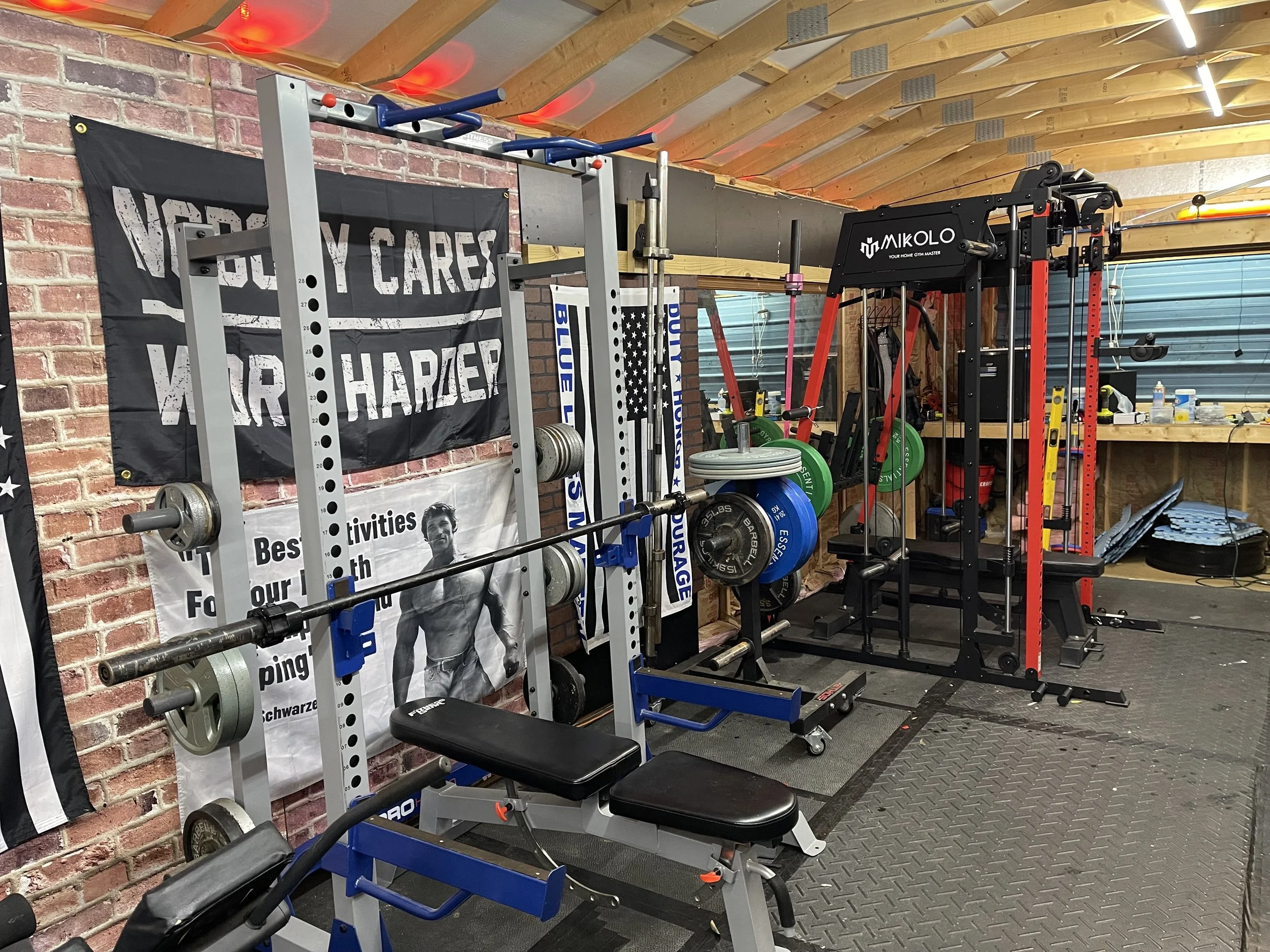 Home gym with weightlifting equipment, barbells, weight plates, a gym bench, and a power rack, with motivational posters and a flag on the brick wall.