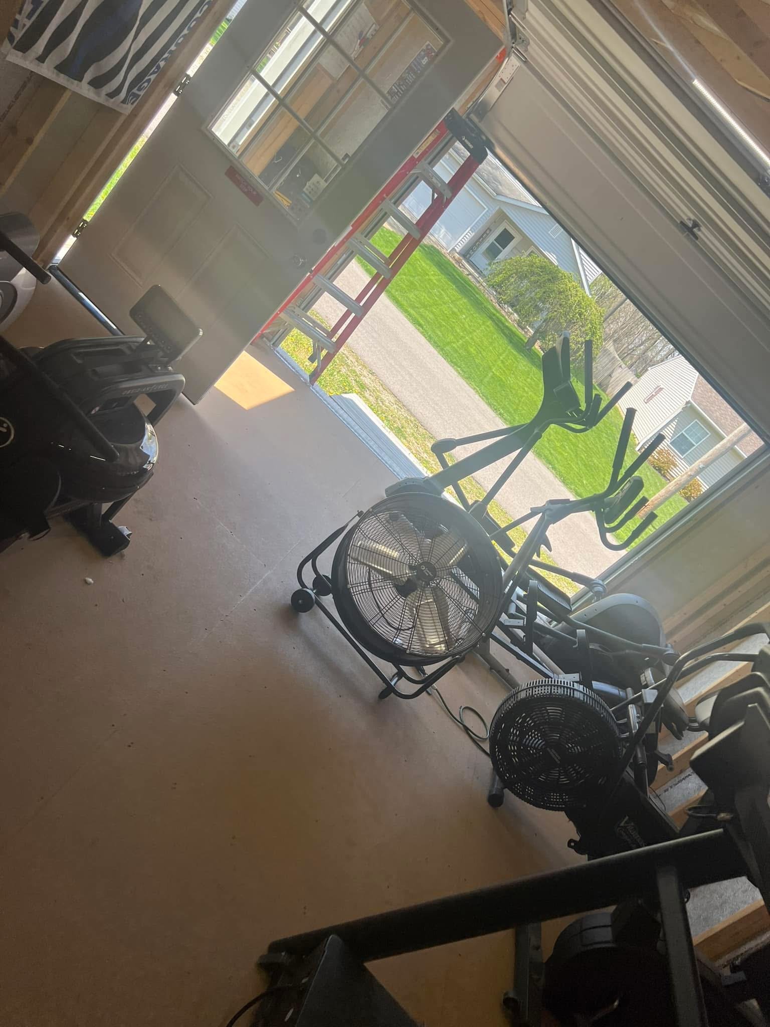 A room with exercise equipment including elliptical machines and fans, with a view of a green lawn and houses outside through large windows.