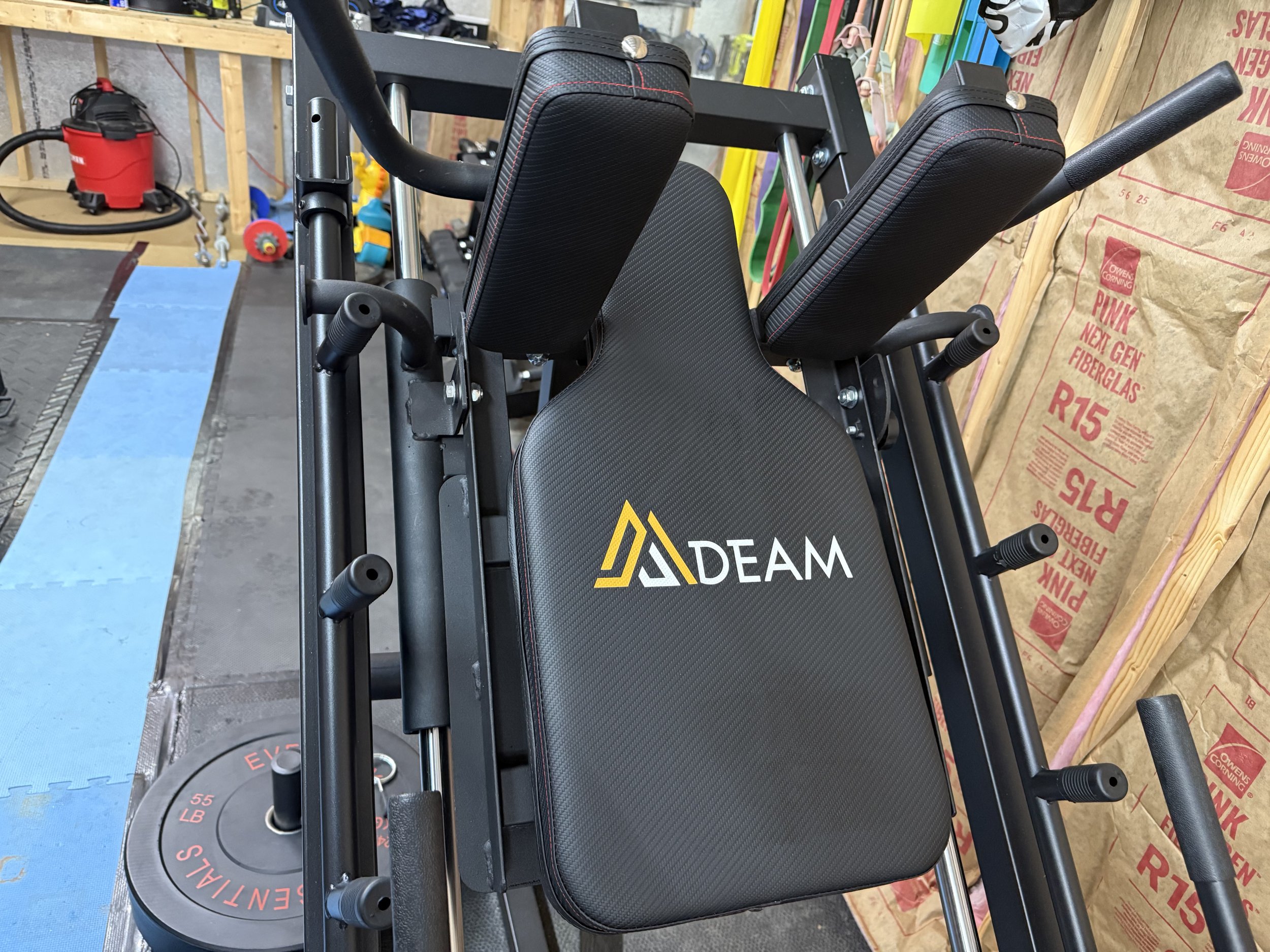 A close-up view of a piece of fitness equipment in a gym, featuring a black padded seat with the logo 'DREAM' in yellow and white, attached to a black metal frame with various padded arm supports.
