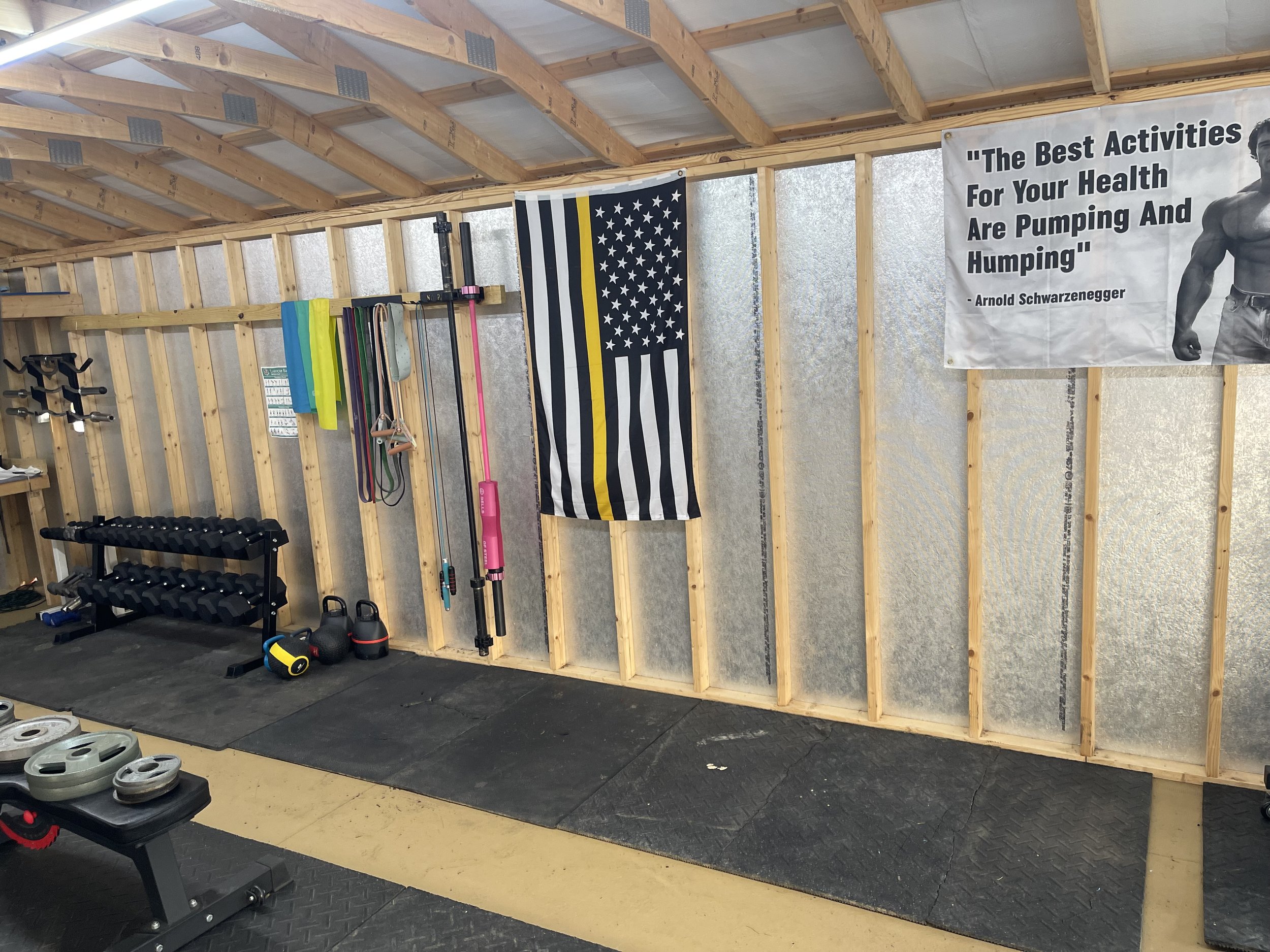 Home gym with weight rack, kettlebells, resistance bands, foam rollers, and a banner with a quote about health activities, next to a black and white American flag and a motivational poster of Arnold Schwarzenegger.