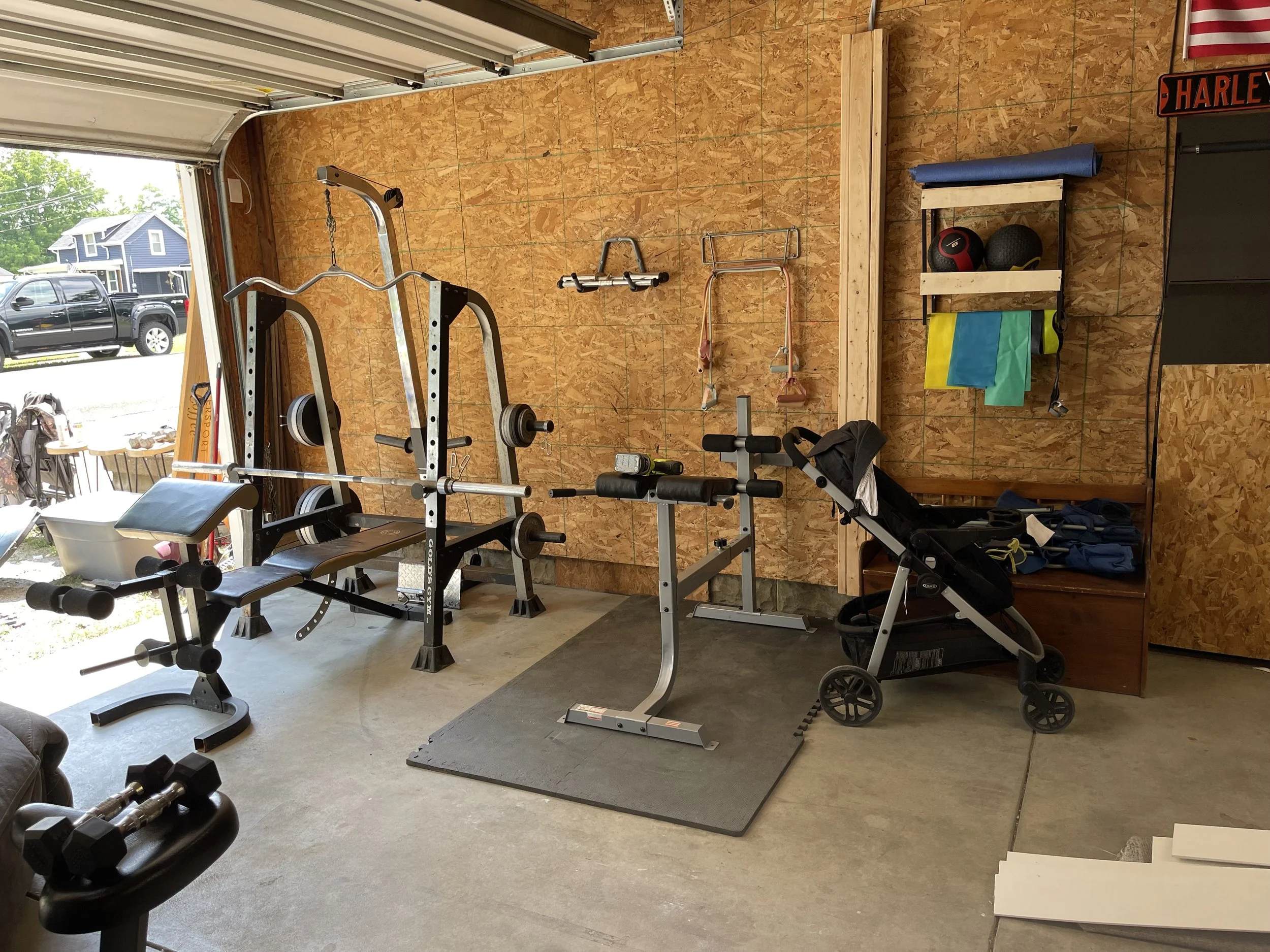 Home gym with weights, workout equipment, and exercise accessories in a garage with plywood walls and an open garage door.