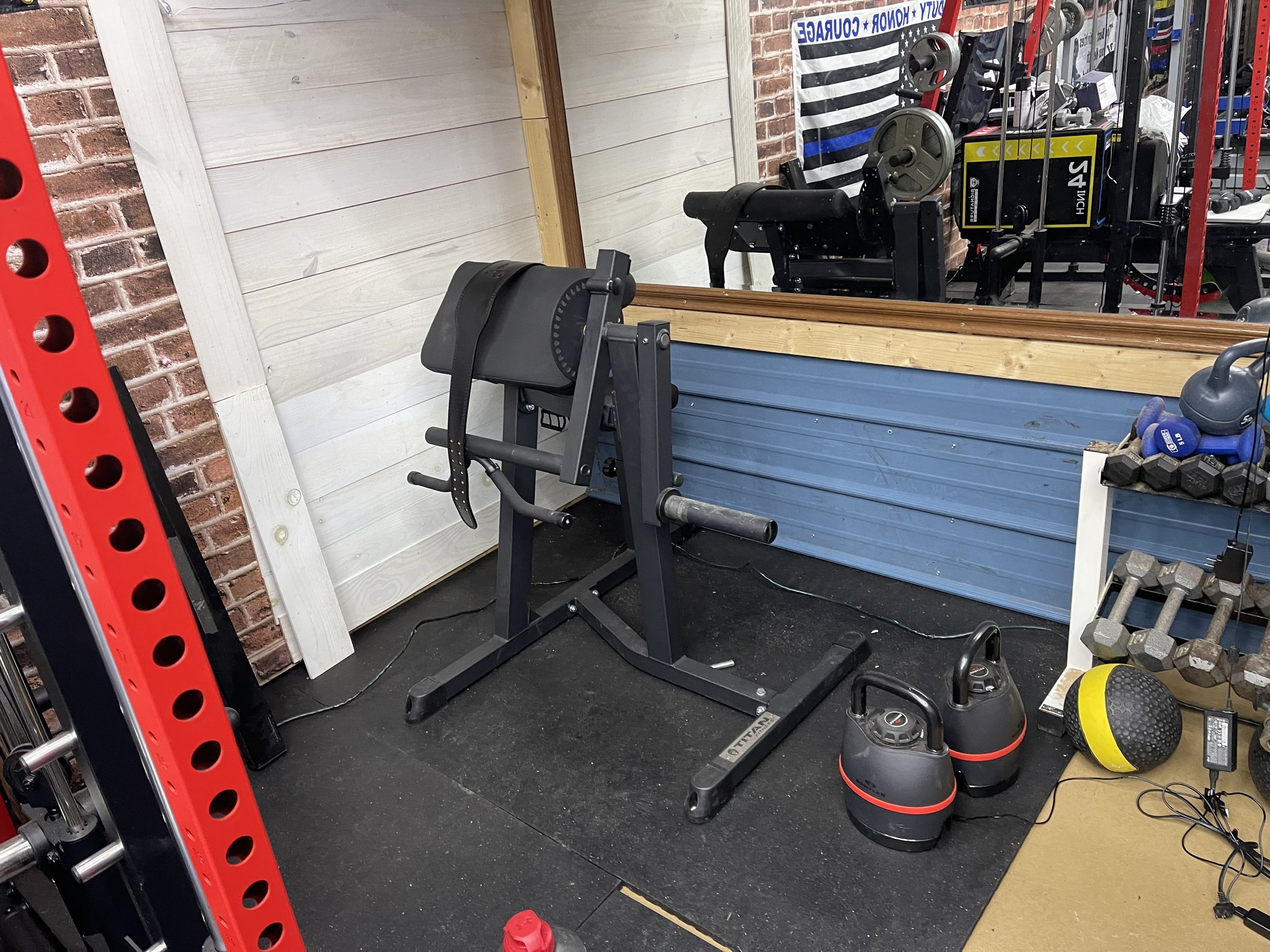 Home gym setup with a workout bench, kettlebells, dumbbells, medicine ball, and weights in a room with brick wall and mirror.
