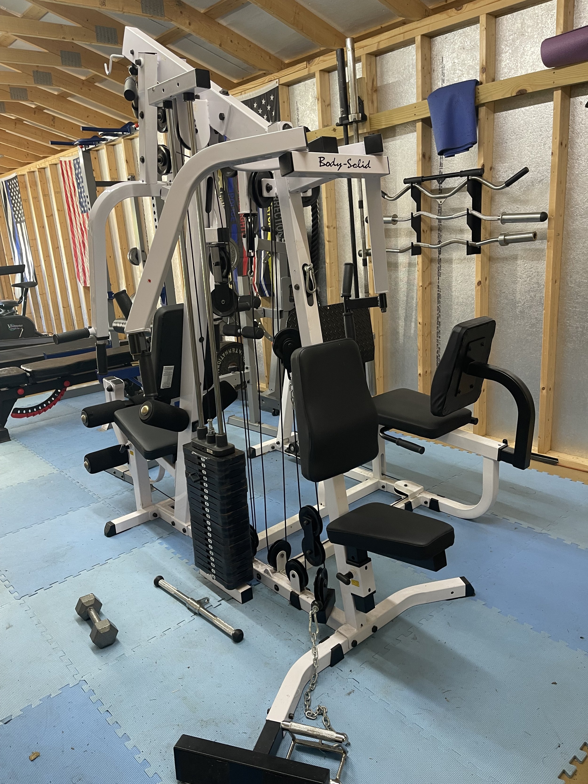 Indoor gym with various exercise equipment including a multi-purpose weight machine, dumbbells, and barbells on a blue padded floor.