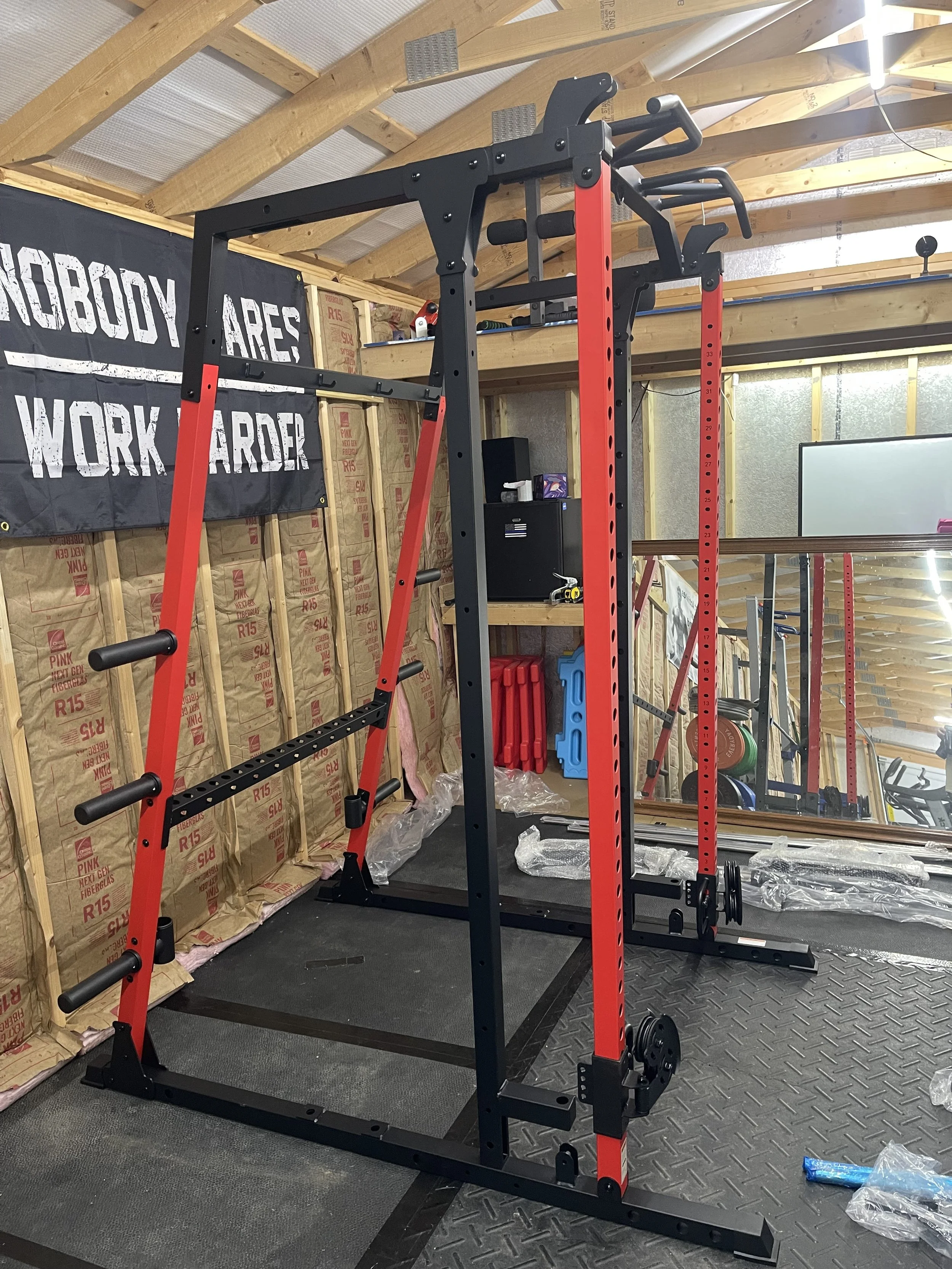 Inside a home gym with a black and red power rack, wooden walls, insulation, and workout equipment including weight plates and a whiteboard.