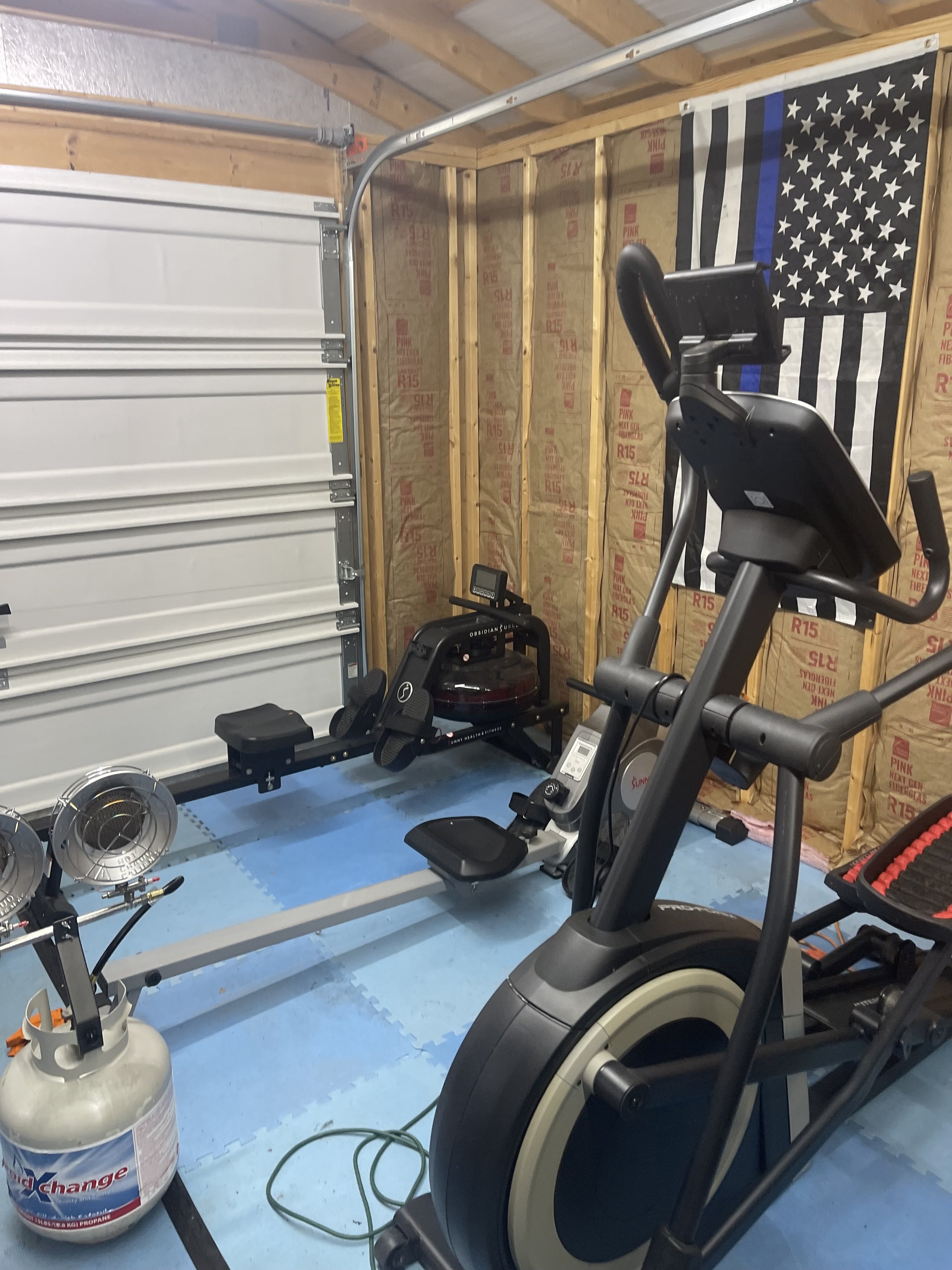 Home gym with exercise bike, rowing machine, and space heater, set up against a wall with a black and white police-themed flag featuring a blue stripe.