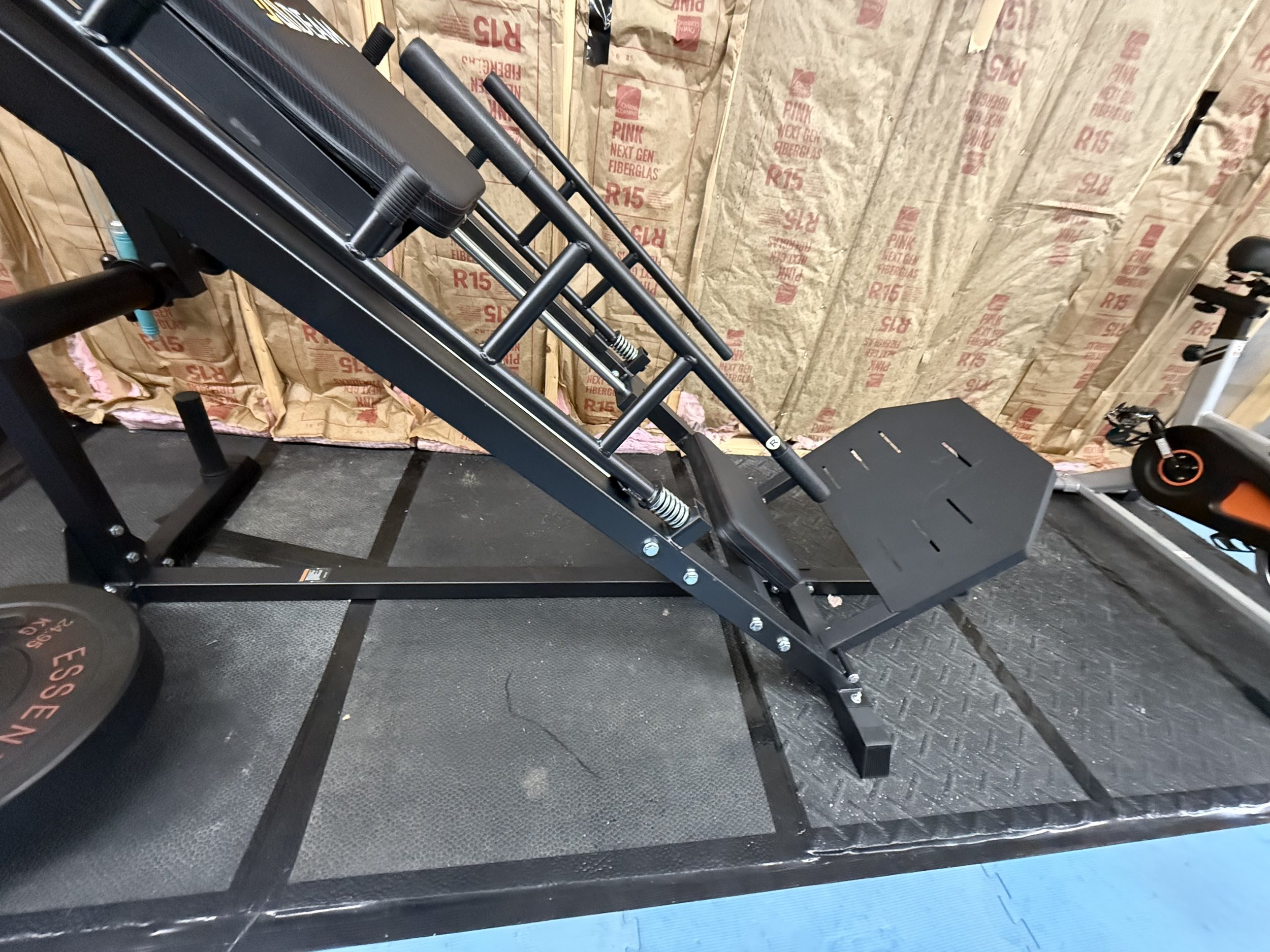 A stair incline trainer machine in a garage gym, with a black frame, adjustable handrails, and a platform for stepping, next to a stationary bike. The backdrop consists of exposed insulation walls.