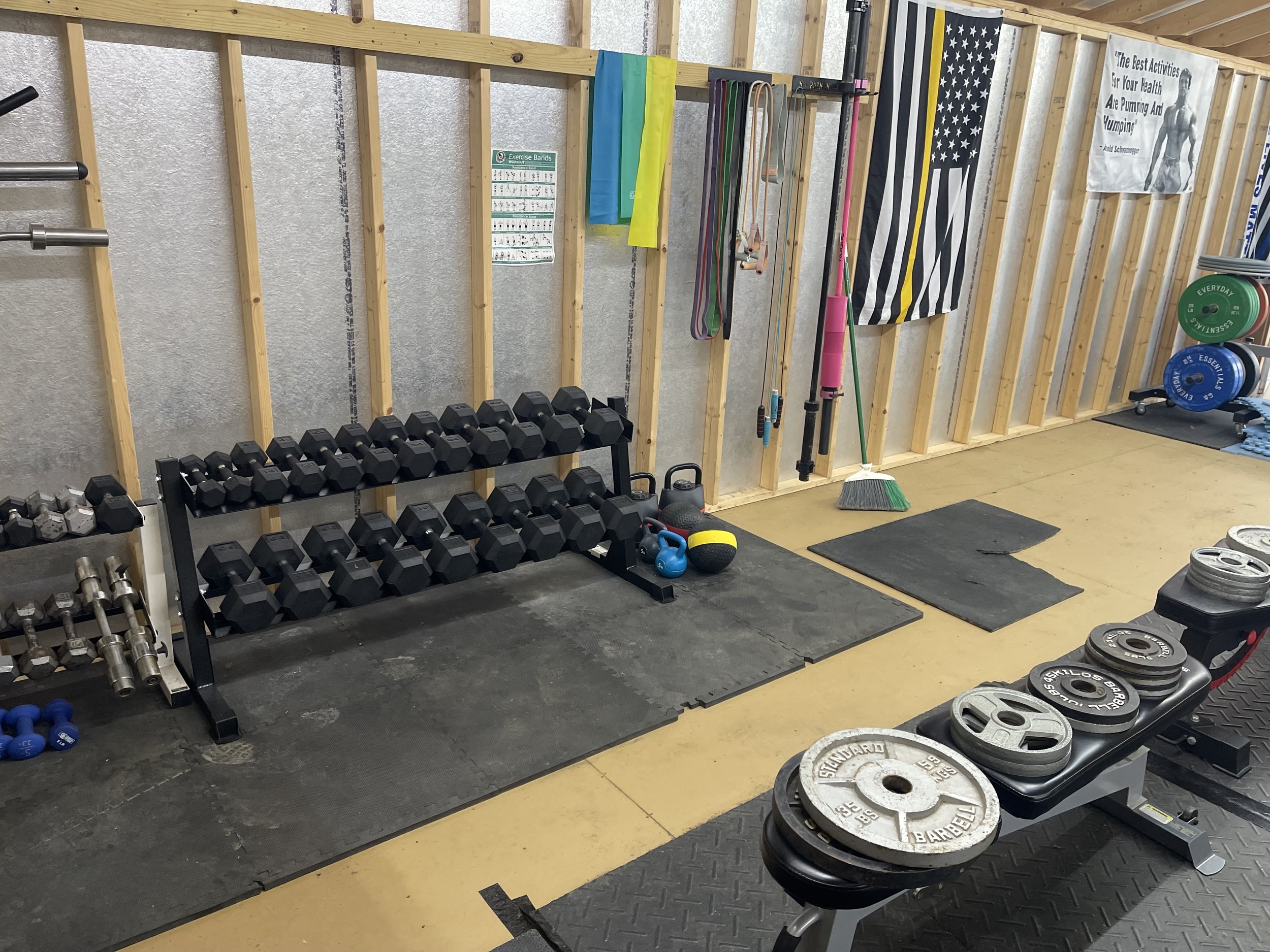 A gym corner with a rack of black dumbbells, kettlebells, a stack of weight plates, and various exercise equipment including resistance bands, jump ropes, and a broom, with motivational poster on the wall.