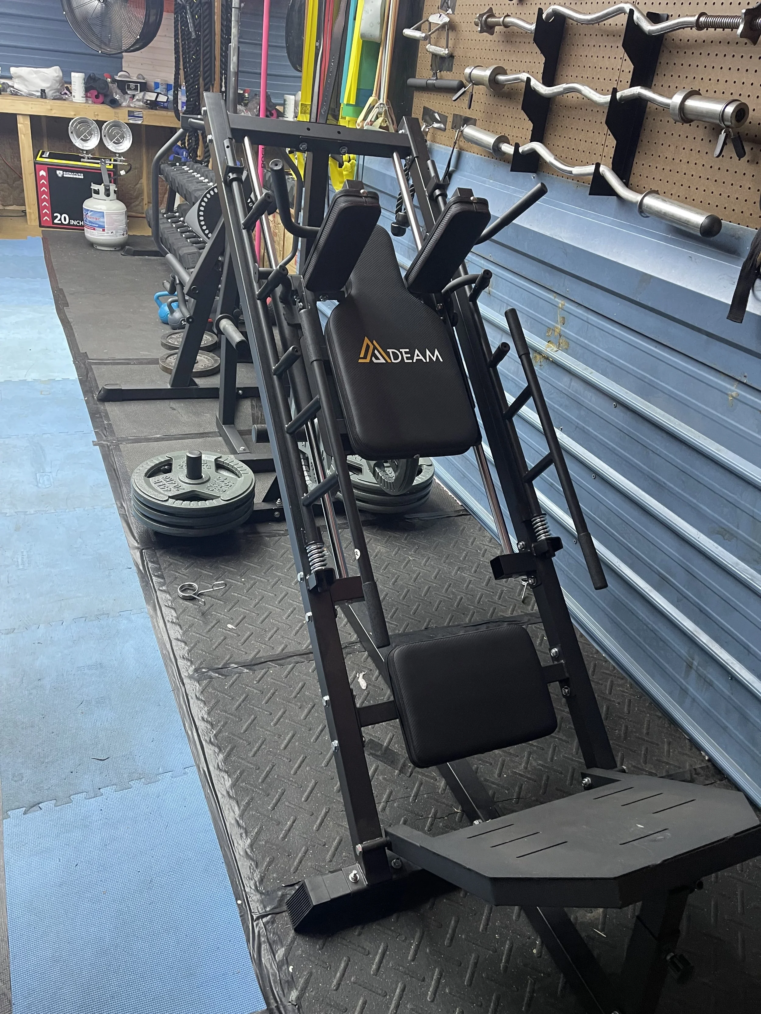 Stationary leg press machine with black padding, used for strength training, in a gym with weight plates, barbells on a pegboard, and various workout equipment in the background.