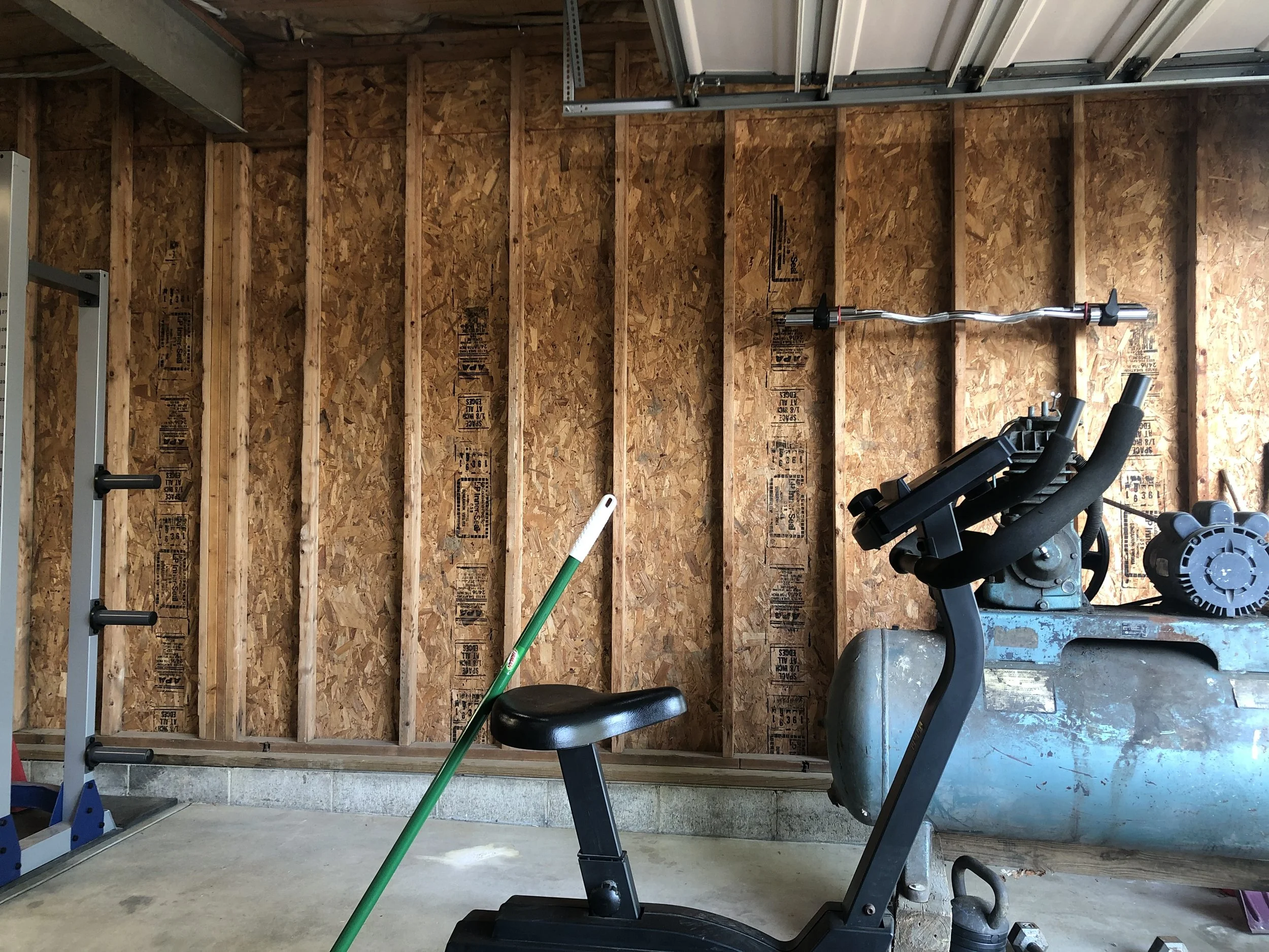 Garage with exposed wooden wall framing, workout equipment including an elliptical machine, a barbell mounted on the wall, and a broom leaning against the elliptical machine.