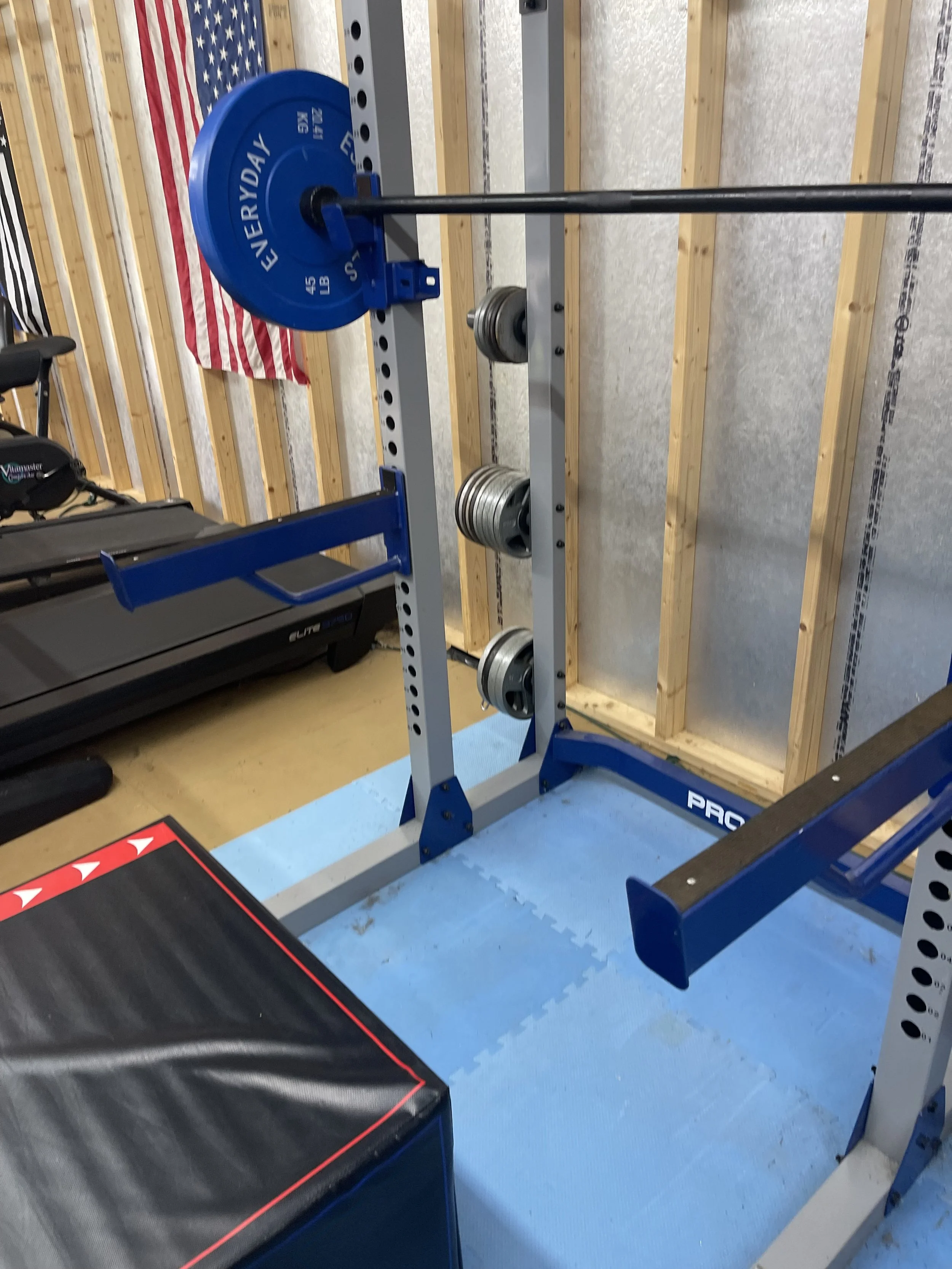 A gym setup with a weightlifting barbell on a squat rack, with a blue weight plate labeled "LIVERY DAY" and several smaller weights on the rack, a treadmill in the background, a black-and-red workbench in the foreground, and an American flag hanging 