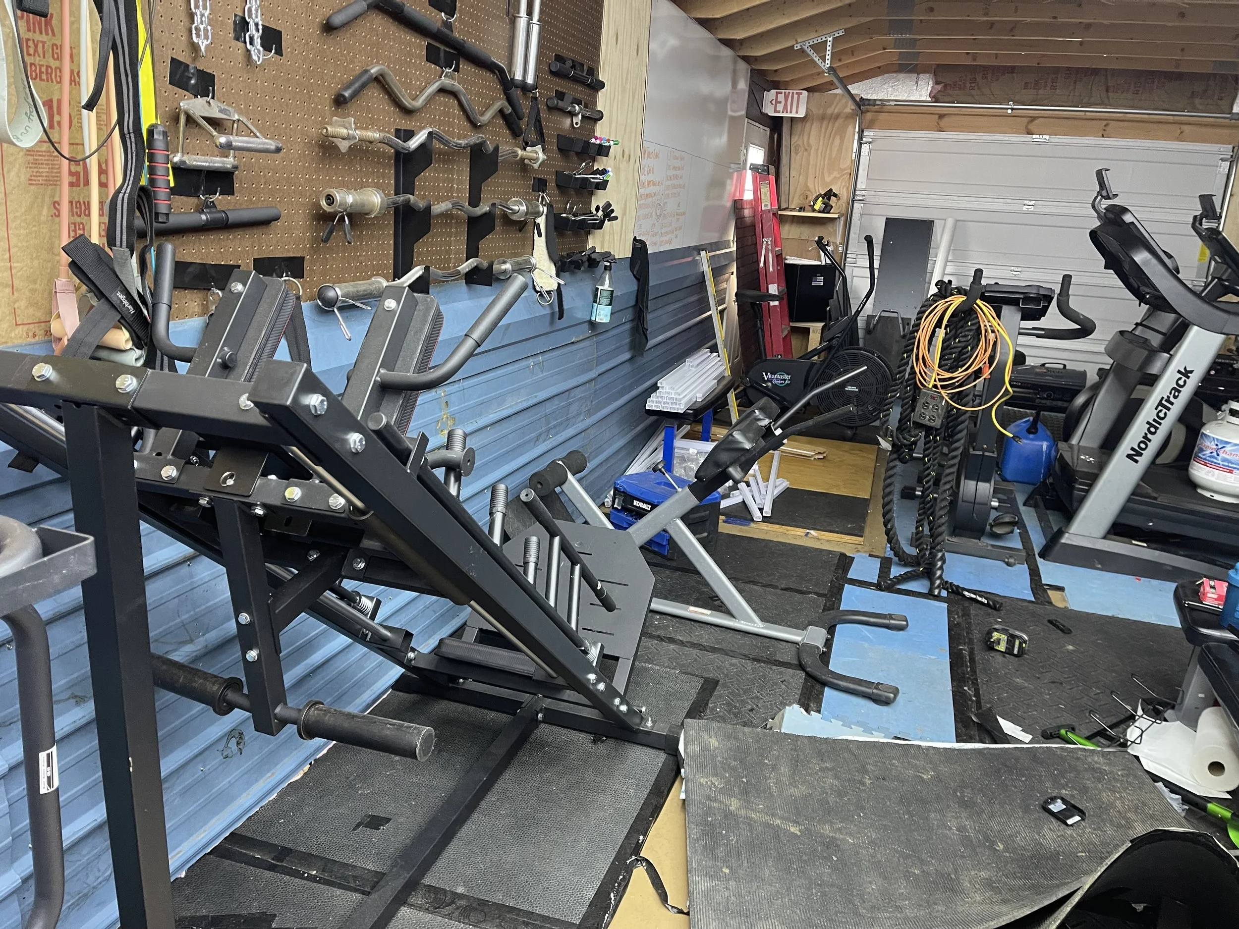 Home gym with various workout equipment including a weight machine, exercisers, a fan, jump ropes, and a pegboard with hand tools on the wall.
