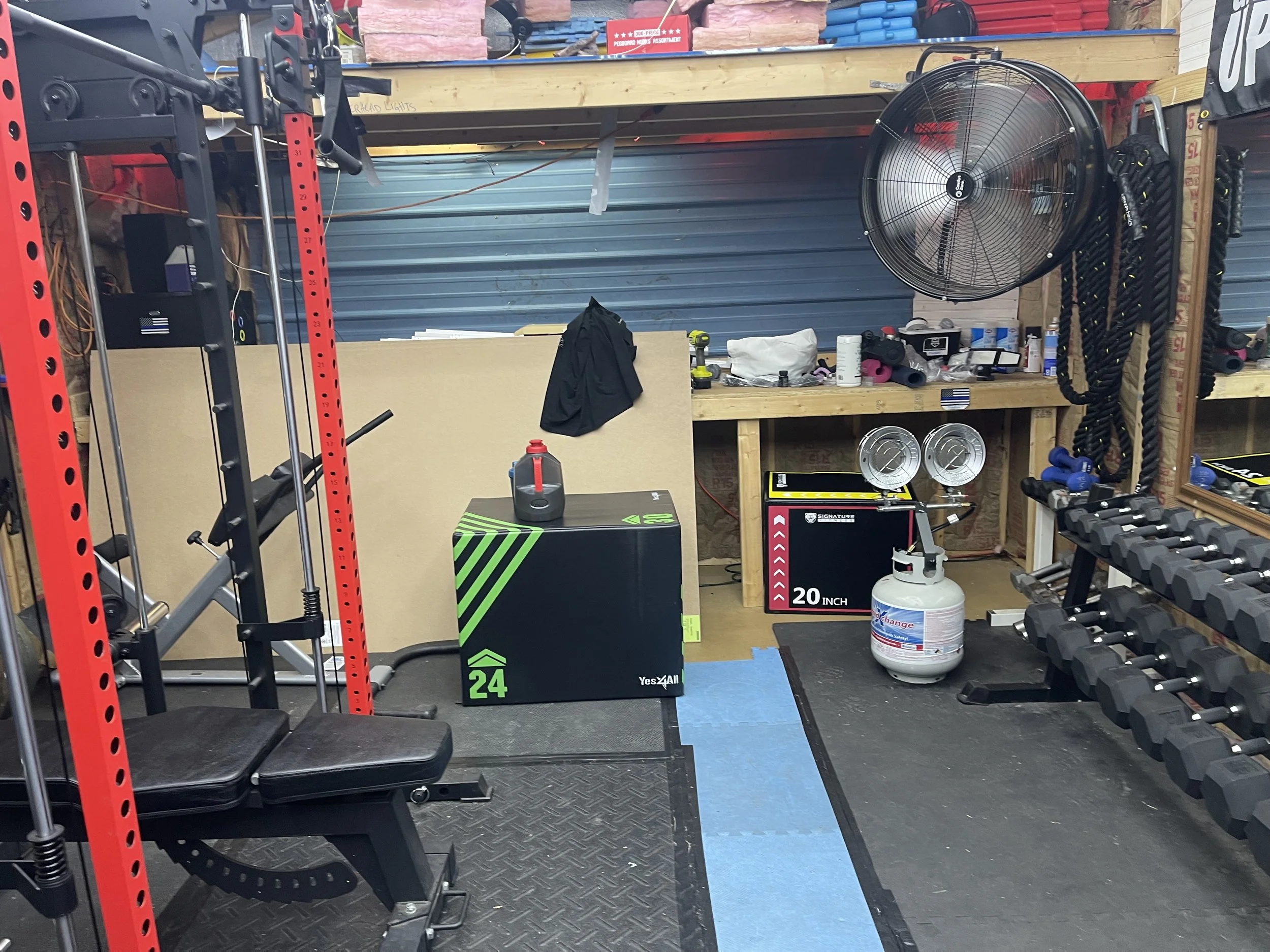 Indoor gym with workout equipment including dumbbells, a bench, a fan, and a propane tank.