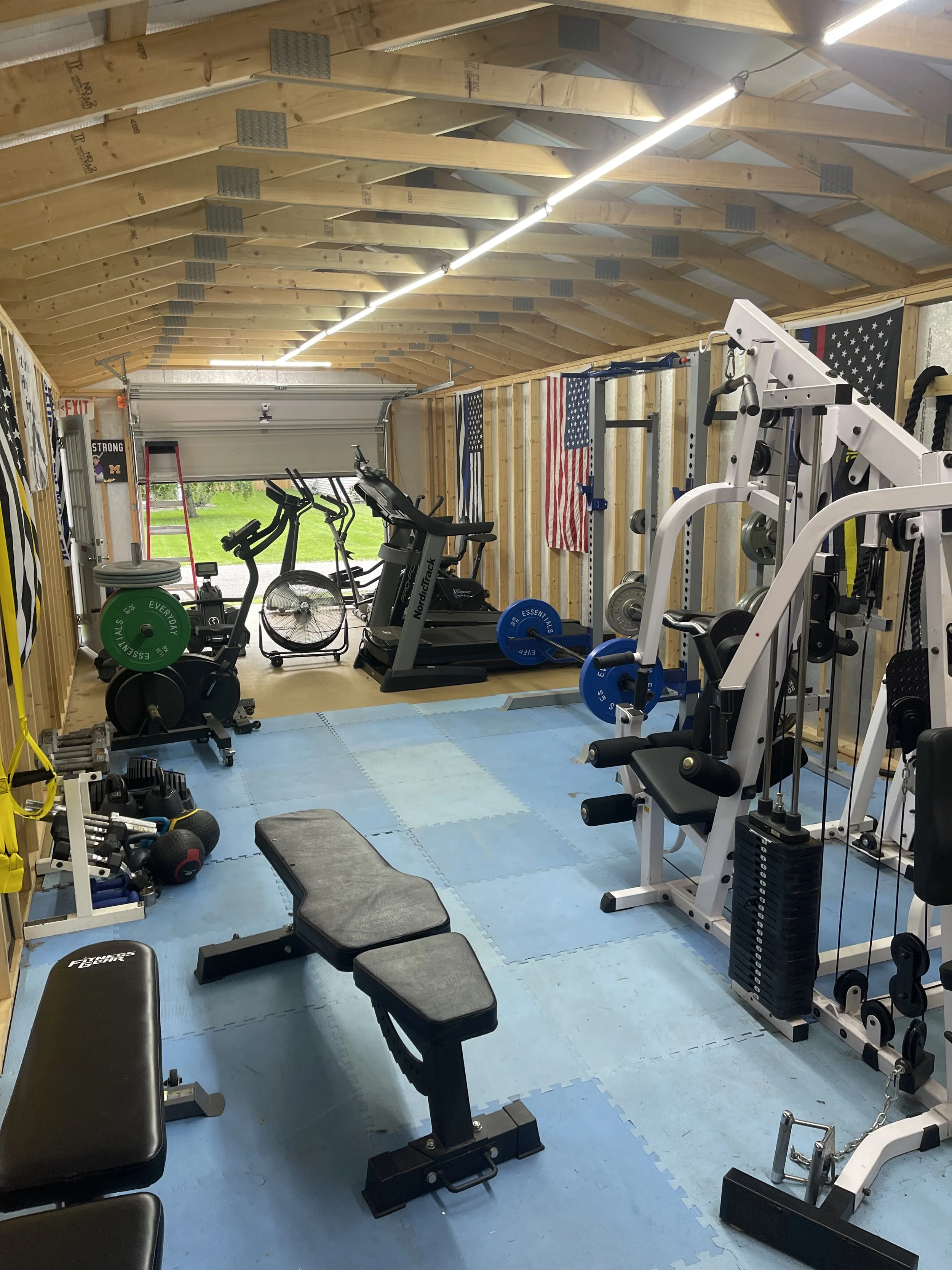 A home gym with various exercise equipment including a weight bench, exercise bike, treadmill, and cable machine. The walls are wood-paneled with American flags, and there is a garage door at the back.