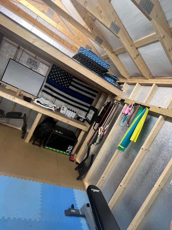Home gym with wooden framing, a flat-screen TV, American flag, folded blue mats, resistance bands, and exercise equipment around a blue foam mat.