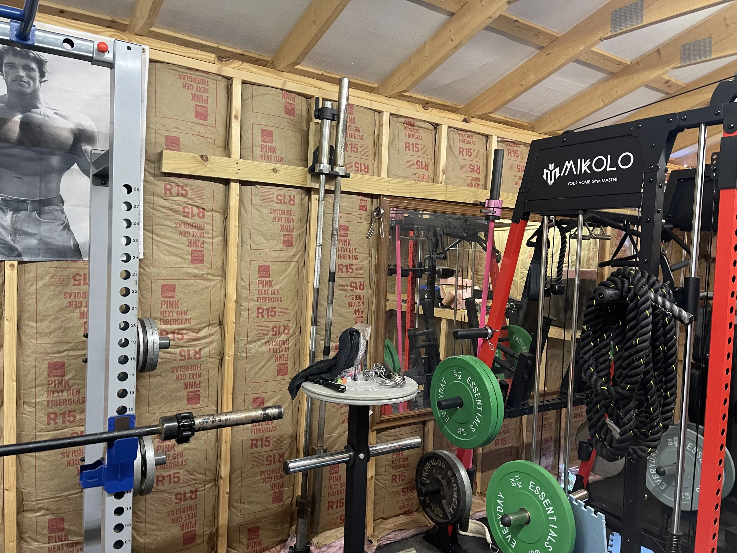 Inside a home gym with various workout equipment including a barbell, weight plates, resistance ropes, and a weight storage rack, against unfinished insulation walls.