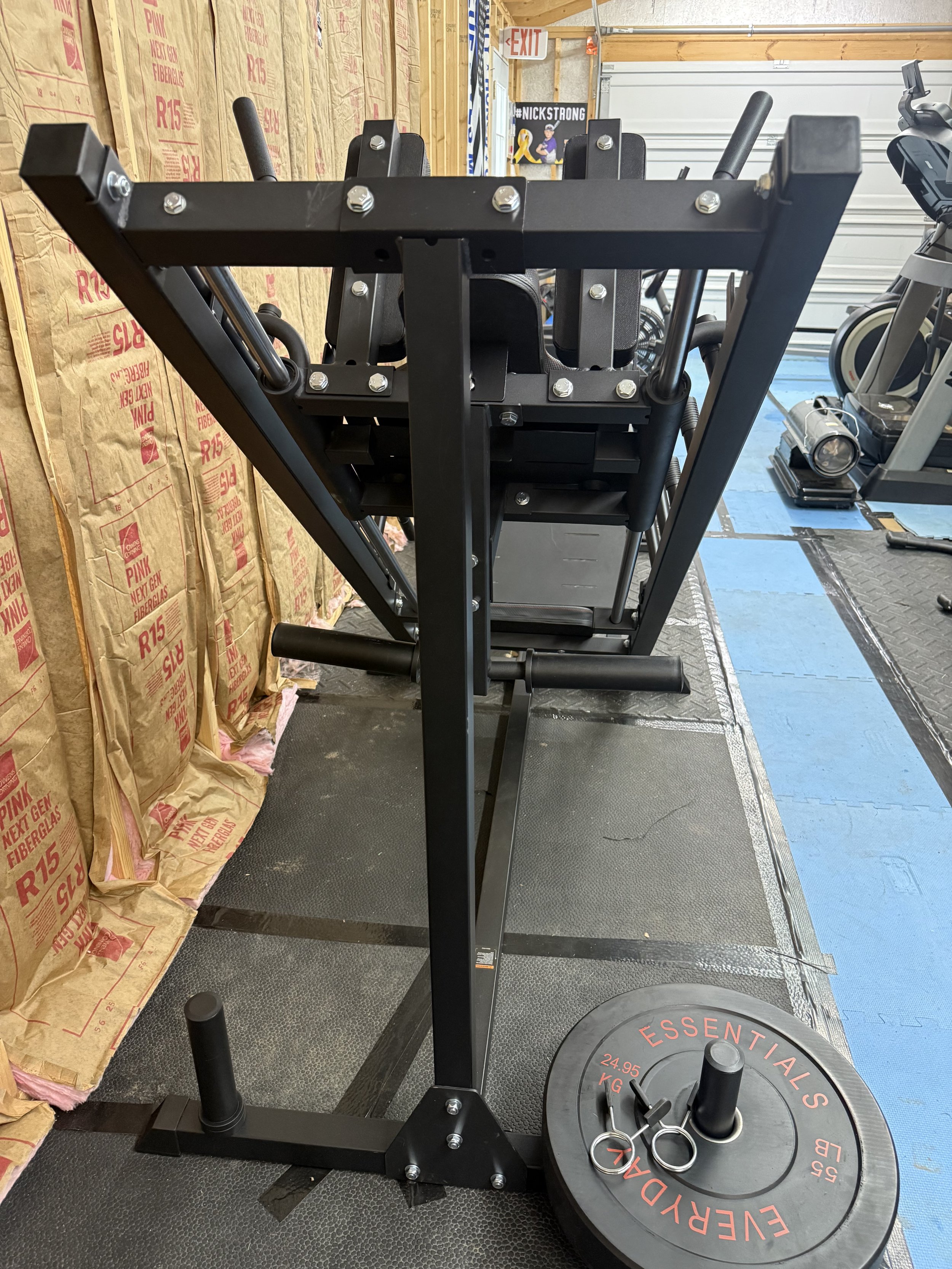 A piece of gym equipment with a weight plate labeled 'Everlast Essentials 24.95 KG' and small pins, in a gym with exercise machines nearby.