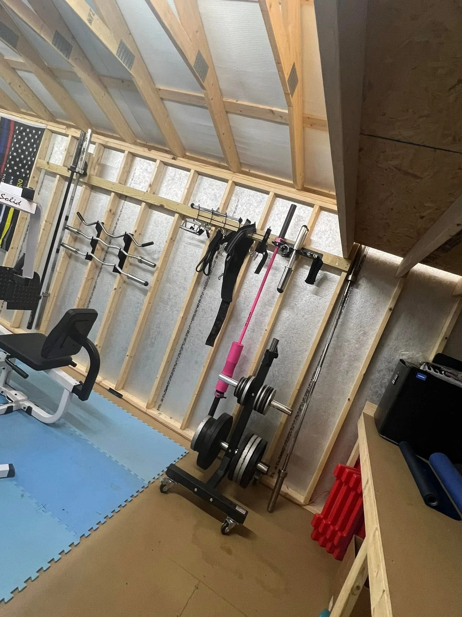 Home gym with weightlifting equipment, dumbbells, a bench, and exercise tools against an unfinished wall with exposed wooden studs.