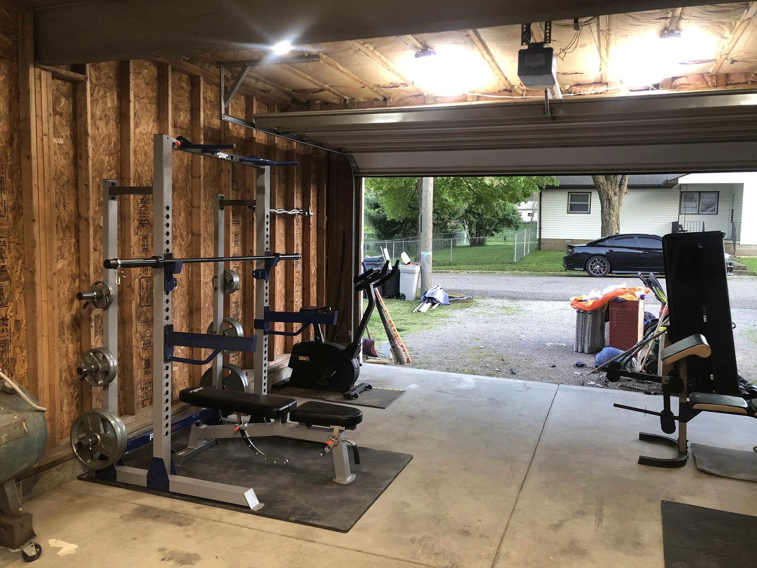 Home gym in a garage with weightlifting equipment, exercise bike, and a view outside of a street with parked cars and a house.