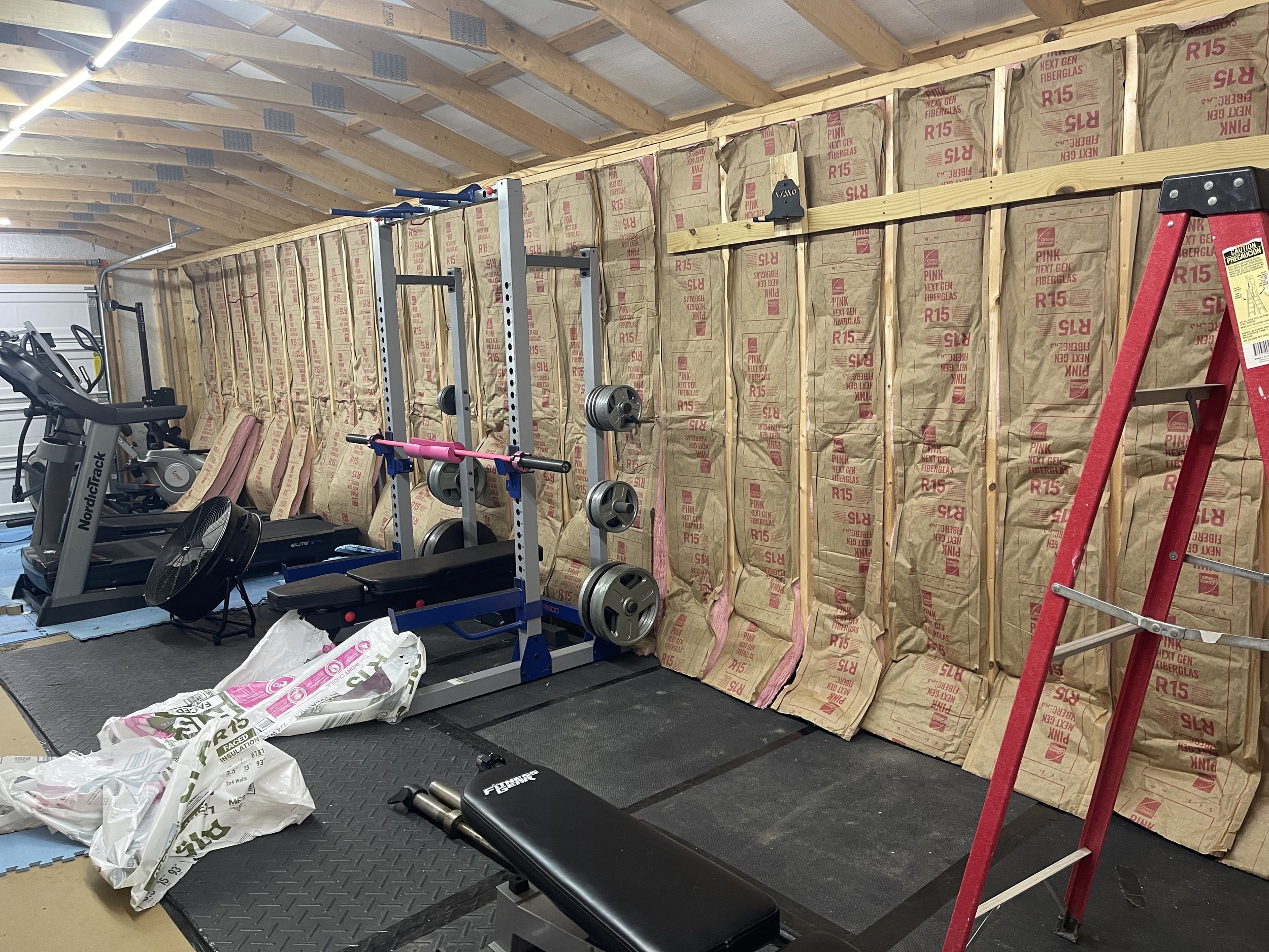 A home gym with workout equipment including a treadmill, a weight rack with plates, a bench, and a red ladder. Insulation is visible on the unfinished wall.