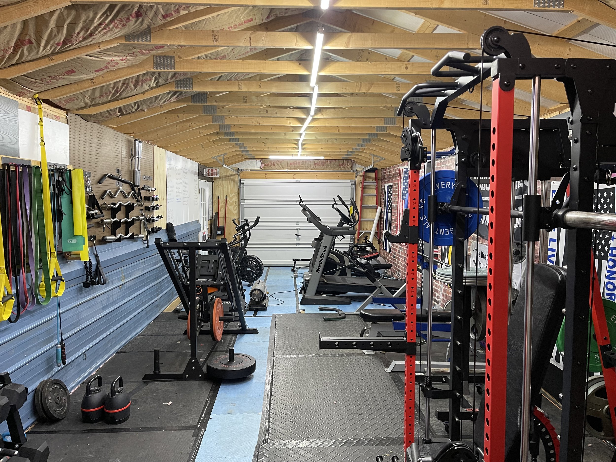 Home gym with various exercise equipment including cardio machines, weightlifting racks, resistance bands, and free weights, inside a garage with plywood walls and a sloped ceiling.