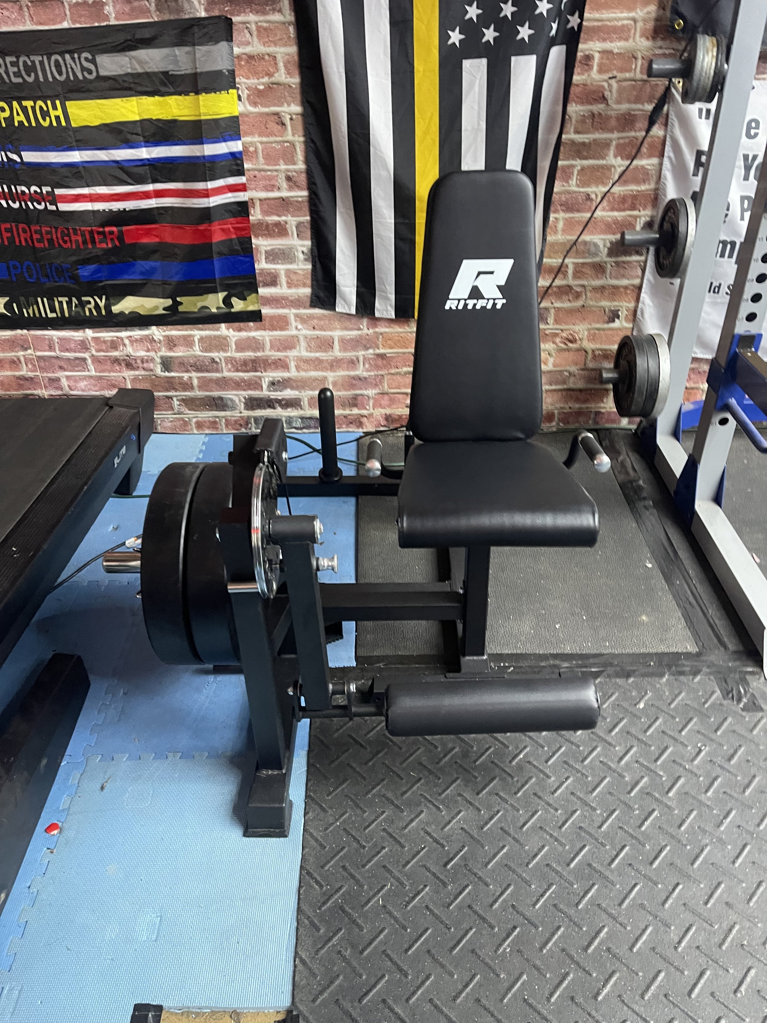 A black seated calf raise machine in a gym, with weight plates on the front, located against a brick wall with posters and flags behind.