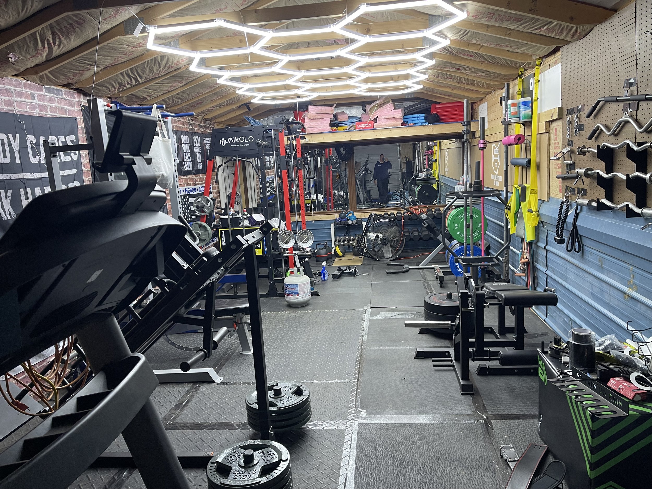 Indoor home gym with various workout equipment, weights, and resistance bands, illuminated by modern hexagonal ceiling lights, with a large mirror on the back wall and a pegboard with fitness accessories on the right.