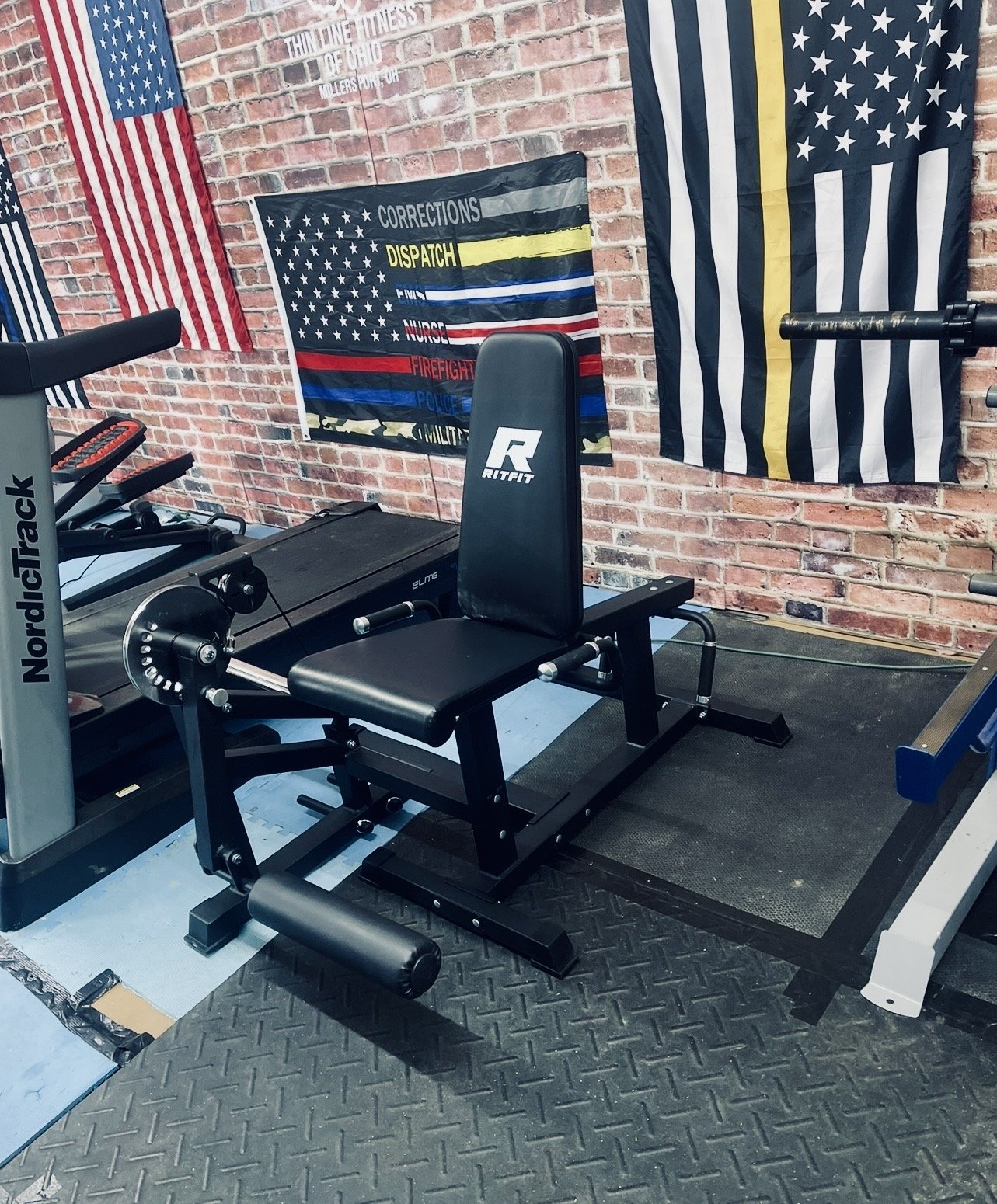 Workout equipment in a gym, including a leg press machine and a treadmill, with American and police flags on a brick wall in the background.
