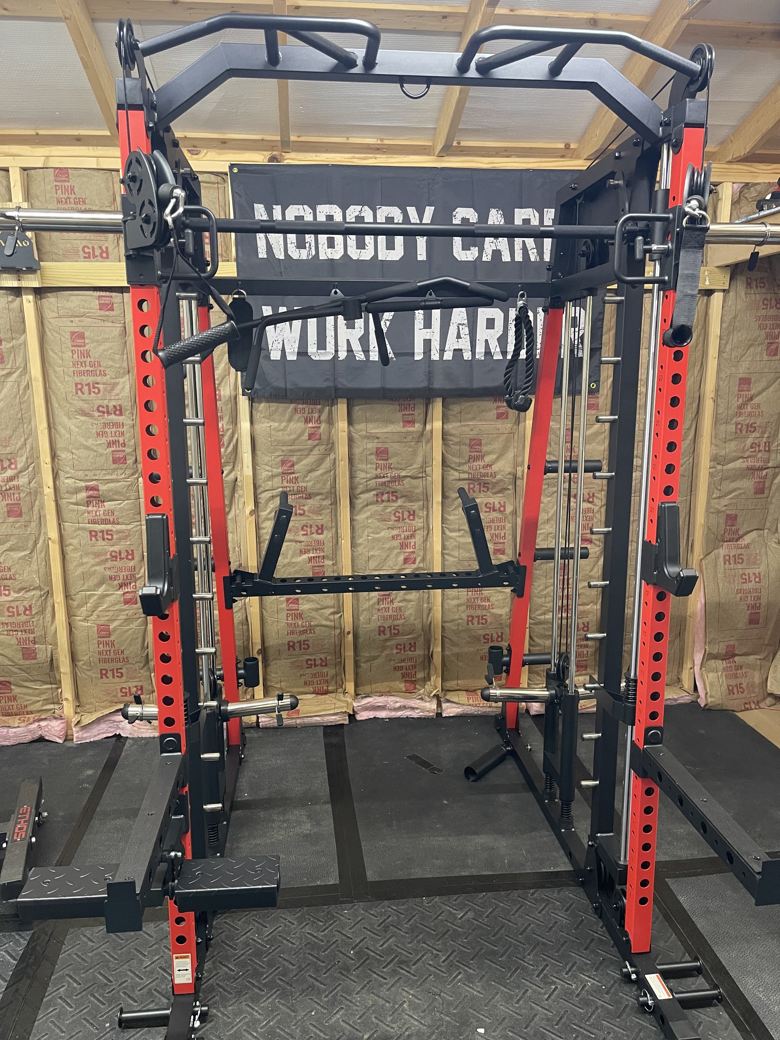 A power rack with various gym equipment mounted, including a pull-up bar, weight storage hooks, and a banner with the text 'Nobody Can Work Hard' hanging in the background.