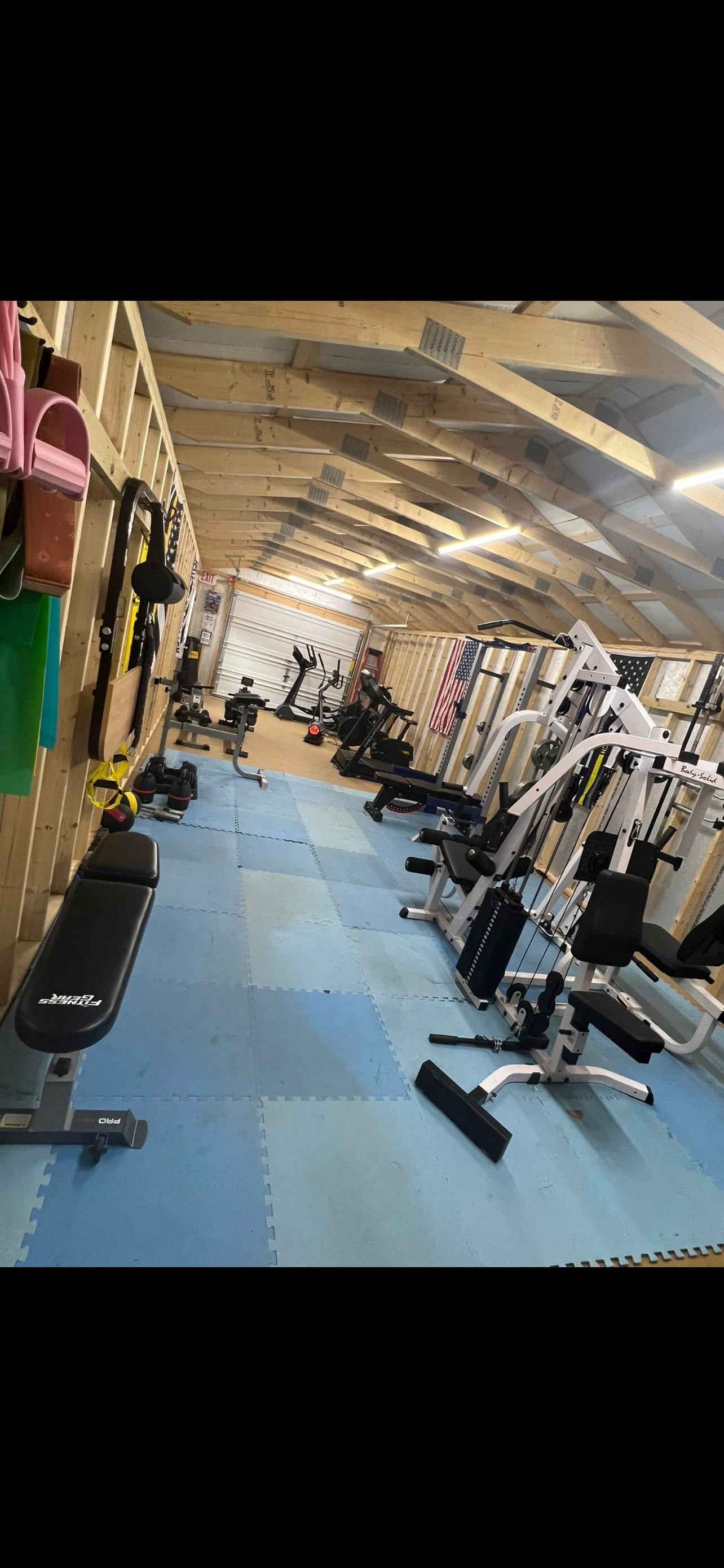 Home gym with various exercise equipment, including weight machines, free weights, and cardio machines, inside a wooden-finished space with an American flag on the wall.