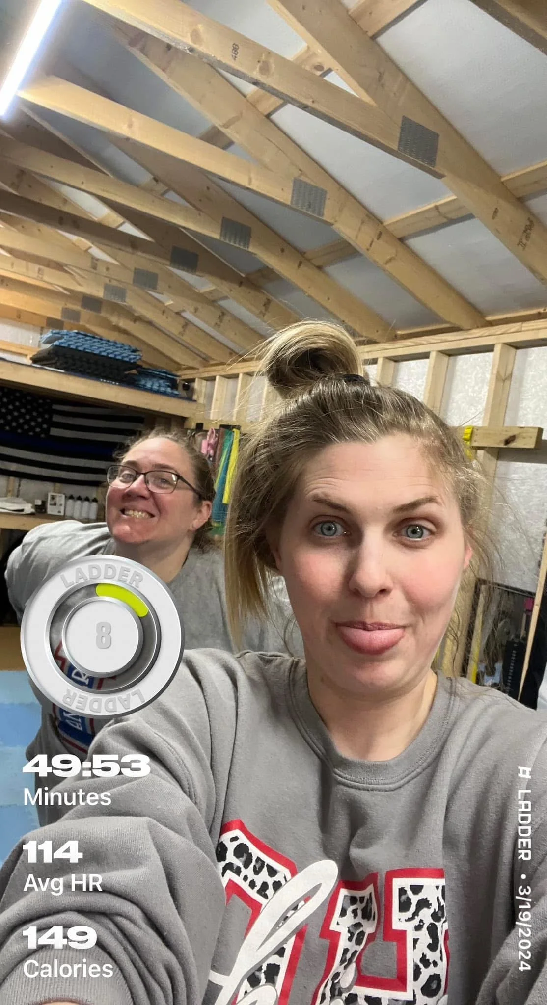Two women taking a selfie inside a partially constructed wooden building or shed. The woman in the foreground is sticking out her tongue, and the woman behind is smiling. Construction tools and an American flag are visible in the background.