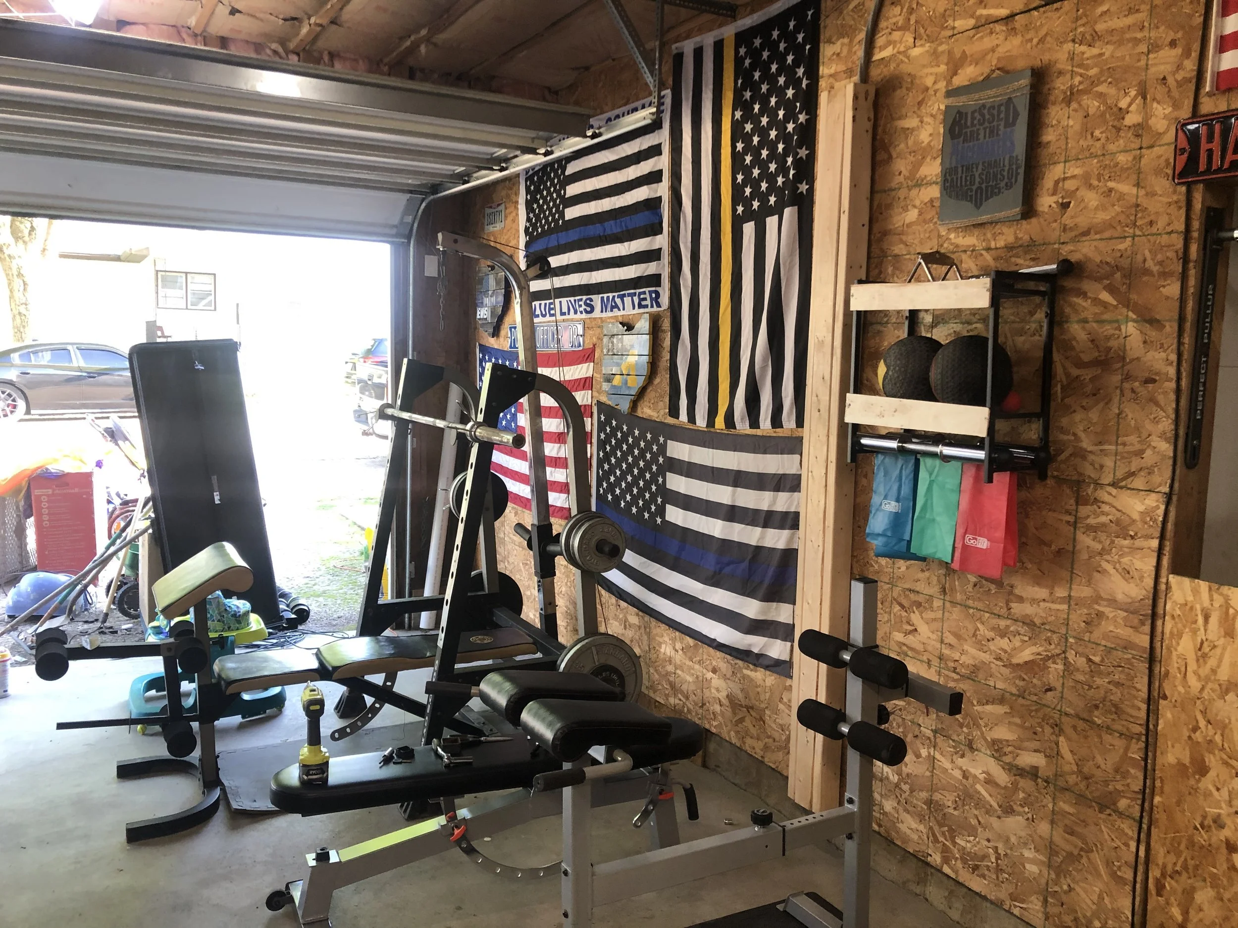 A garage gym with workout equipment, American flags, and patriotic banners on a plywood wall. There are weights, resistance bands, a rowing machine, and a black exercise bench, with sunlight coming in from outside.