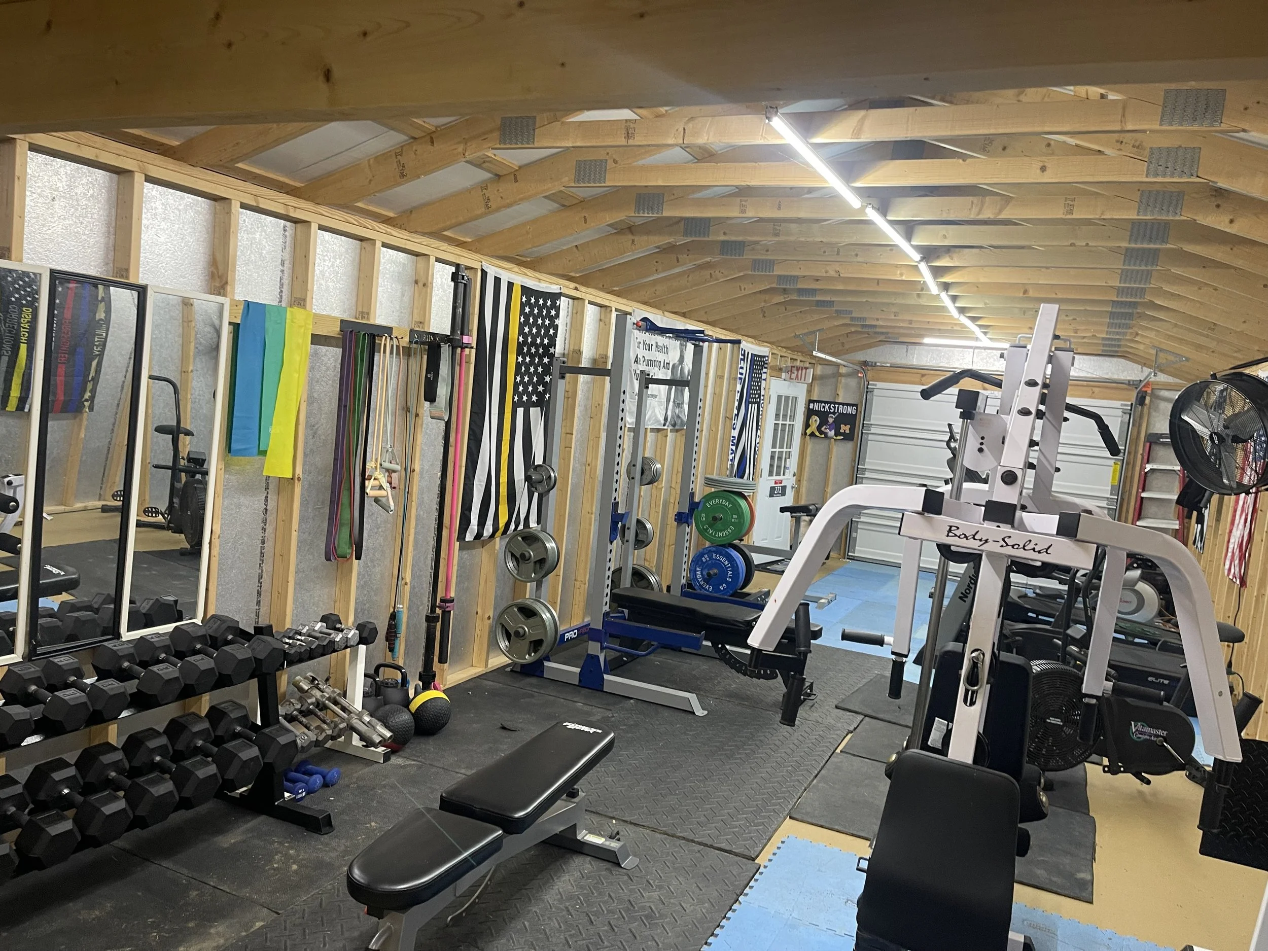 A home gym with various exercise equipment, including dumbbells, weight plates, medicine balls, resistance bands, a bench, and a squat rack. The room has wooden walls and ceiling, rubber flooring, and flags hanging on the wall.