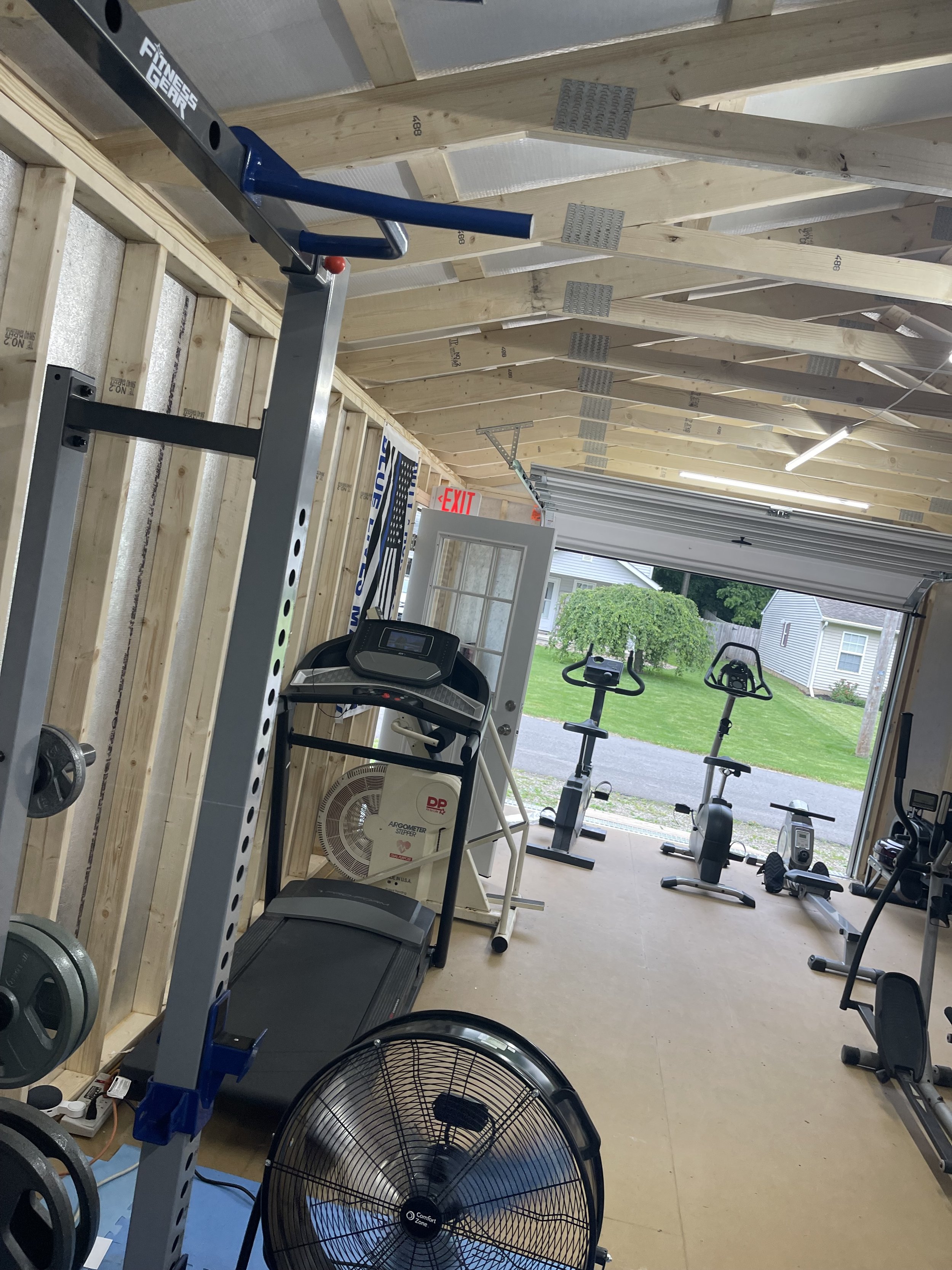 Home gym with treadmill, exercise bikes, fan, and wooden walls with parking lot and houses visible outside.
