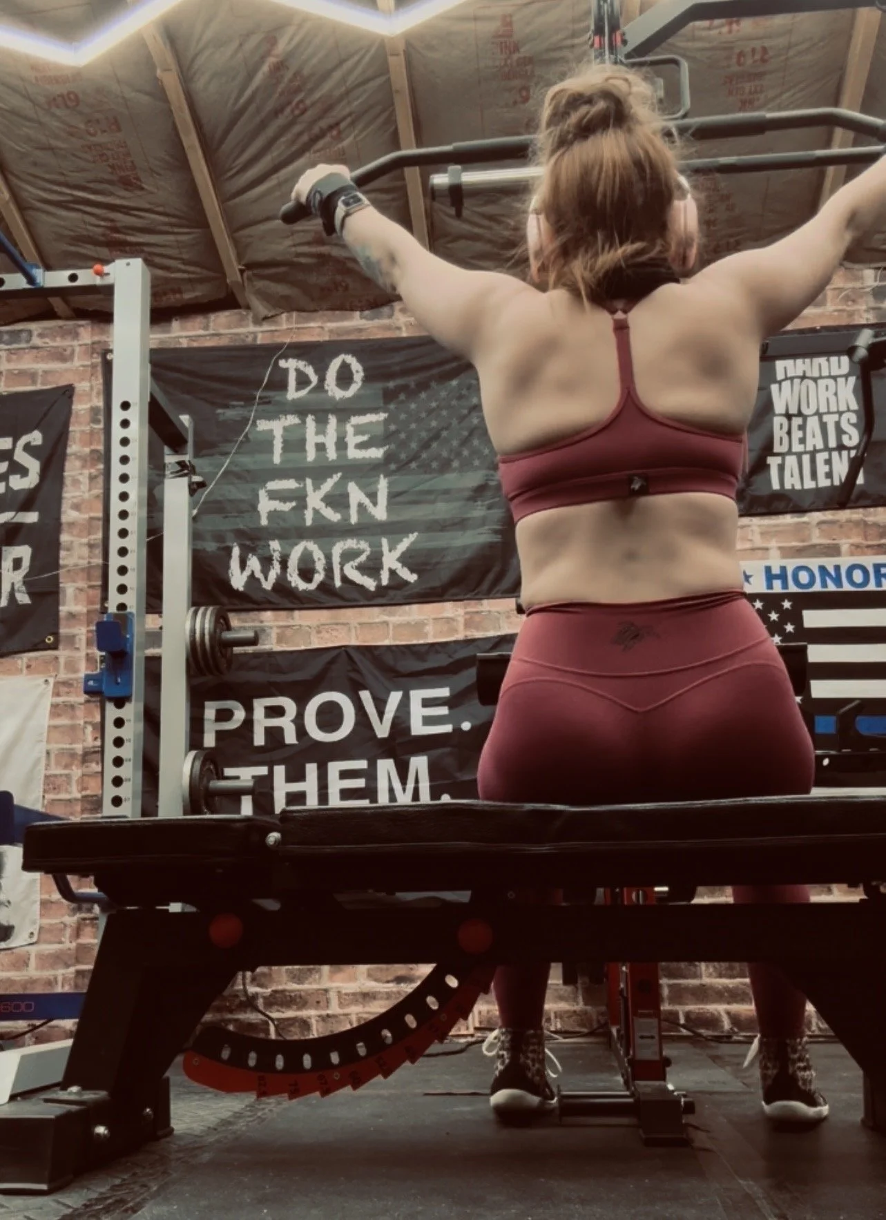 A woman in maroon athletic wear lifting her arms above her head in a gym with motivational banners reading 'Do the FKN work' and 'Prove them.' The gym has exposed brick walls and weightlifting equipment.