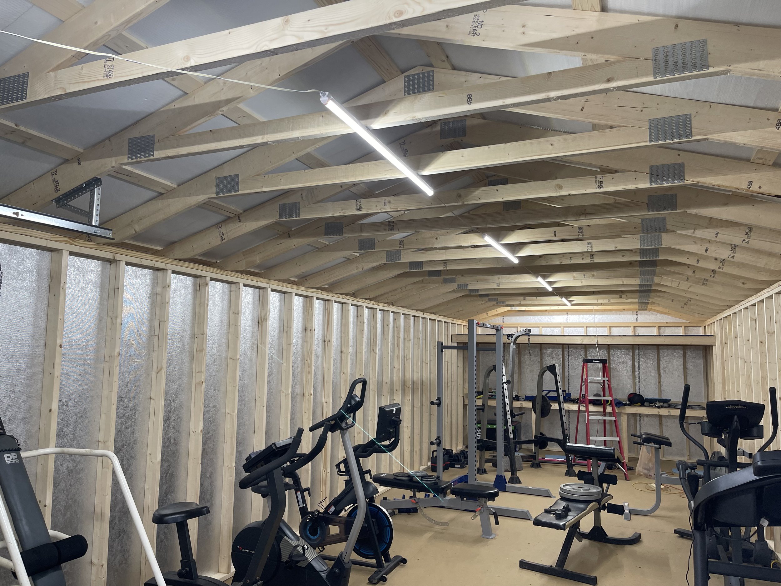 A partially constructed gym or workout space with exposed wooden framing on the walls and ceiling. The ceiling has wooden trusses with metal plates, and the space is lit by long fluorescent lights. Exercise equipment such as stationary bikes, weight 