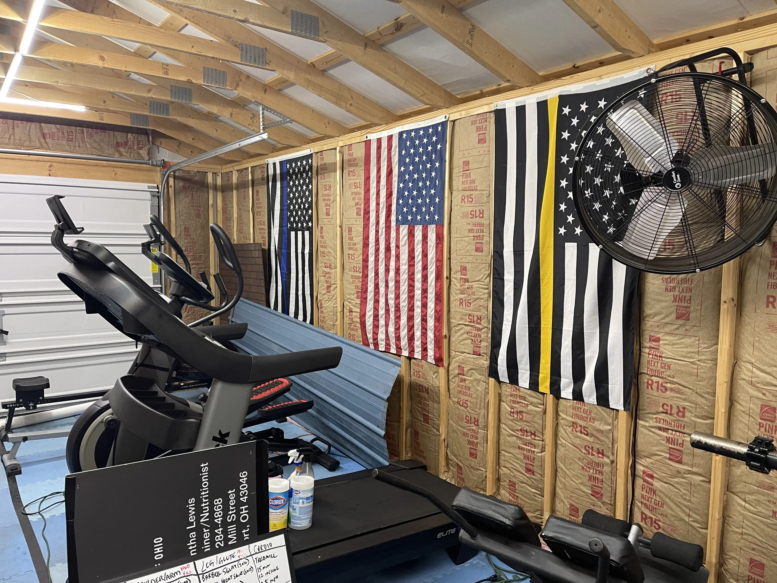 A home gym setup in a garage with an exercise bike, a treadmill, and a rowing machine. The walls are unfinished with exposed insulation and wooden framing. Three American flags and one with a black and white design with a yellow stripe are hanging on
