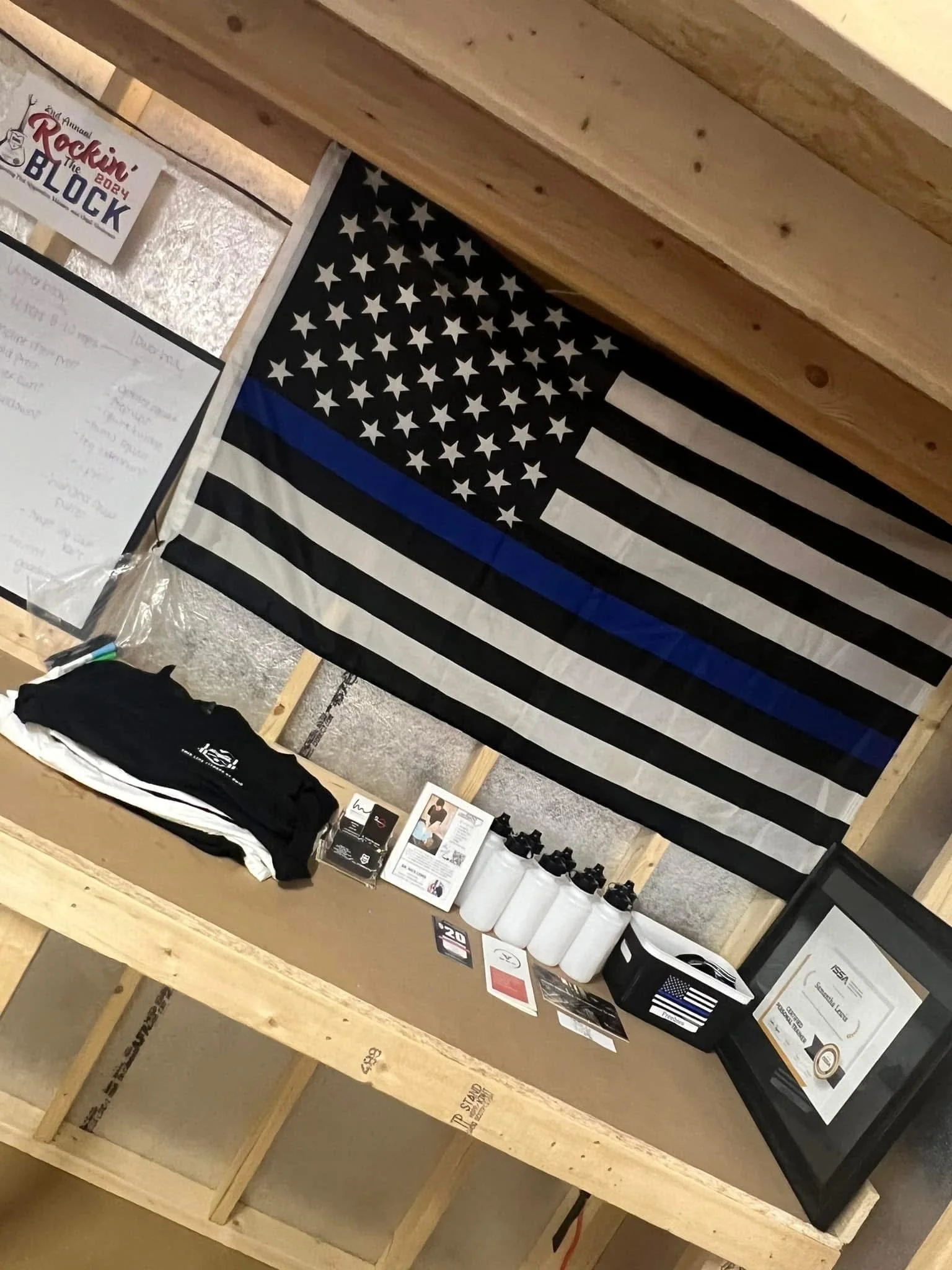Display table with white water bottles, framed certificate, folded clothing, brochures, and business cards in front of a black and white American flag with a blue stripe.