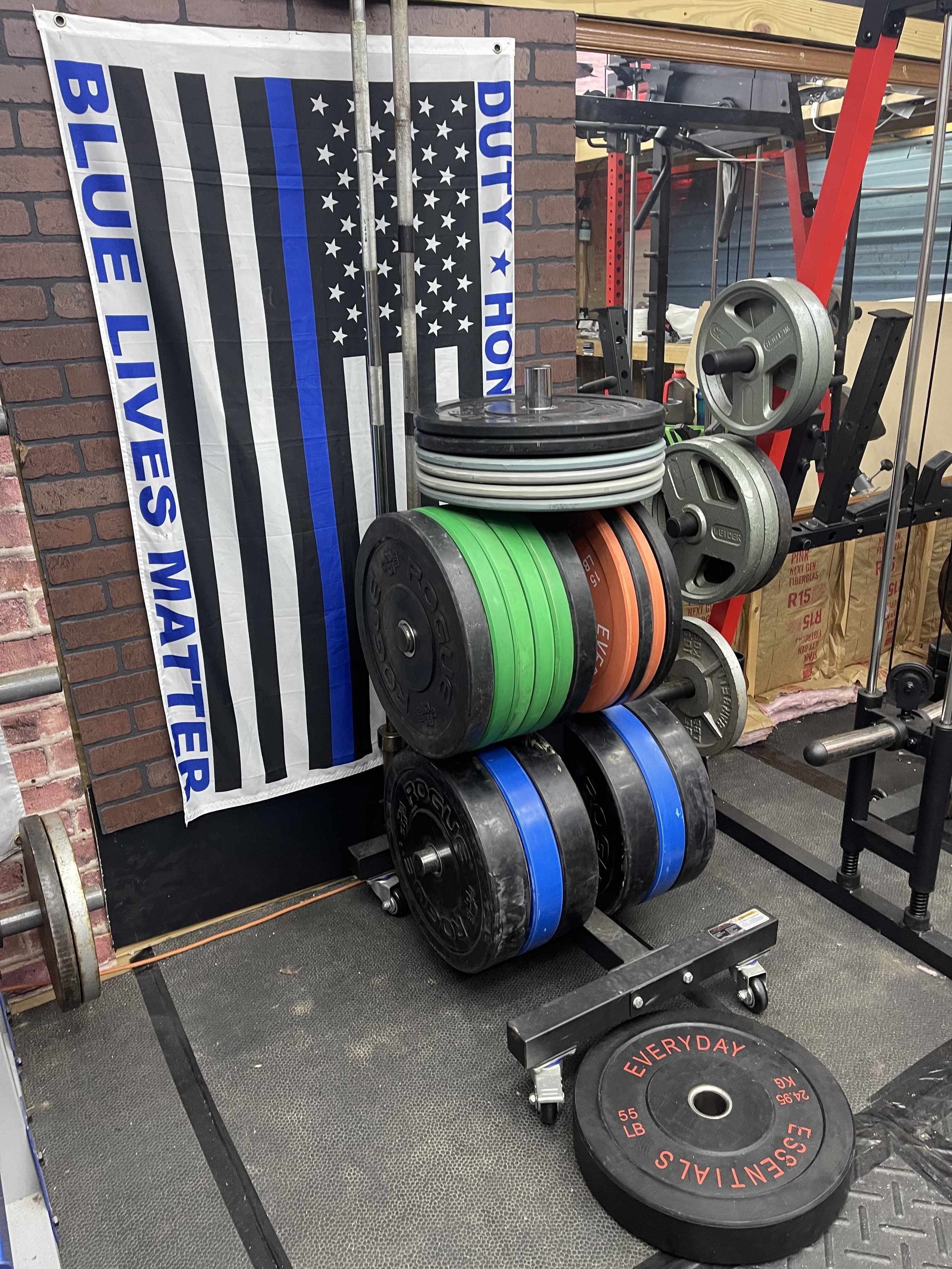 A gym corner with weight plates stacked on a rack, a blue 'Blue Lives Matter' flag with a police badge design and police flag pattern, and gym equipment including a barbell, dumbbells, and weight plates.