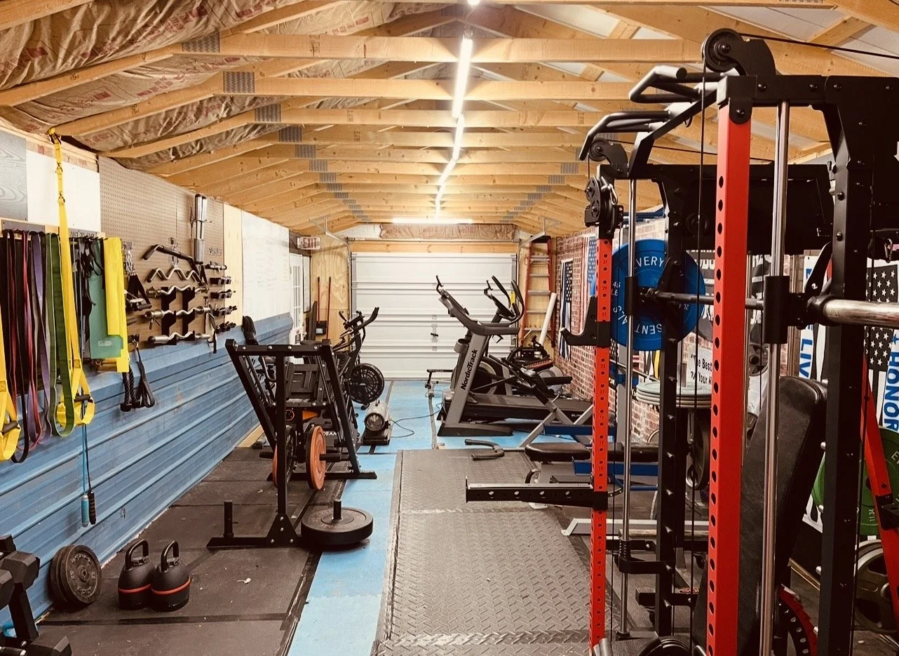 A home gym with various exercise equipment, including treadmills, weights, resistance bands, and other workout gear in a garage with unfinished walls and a wooden ceiling.