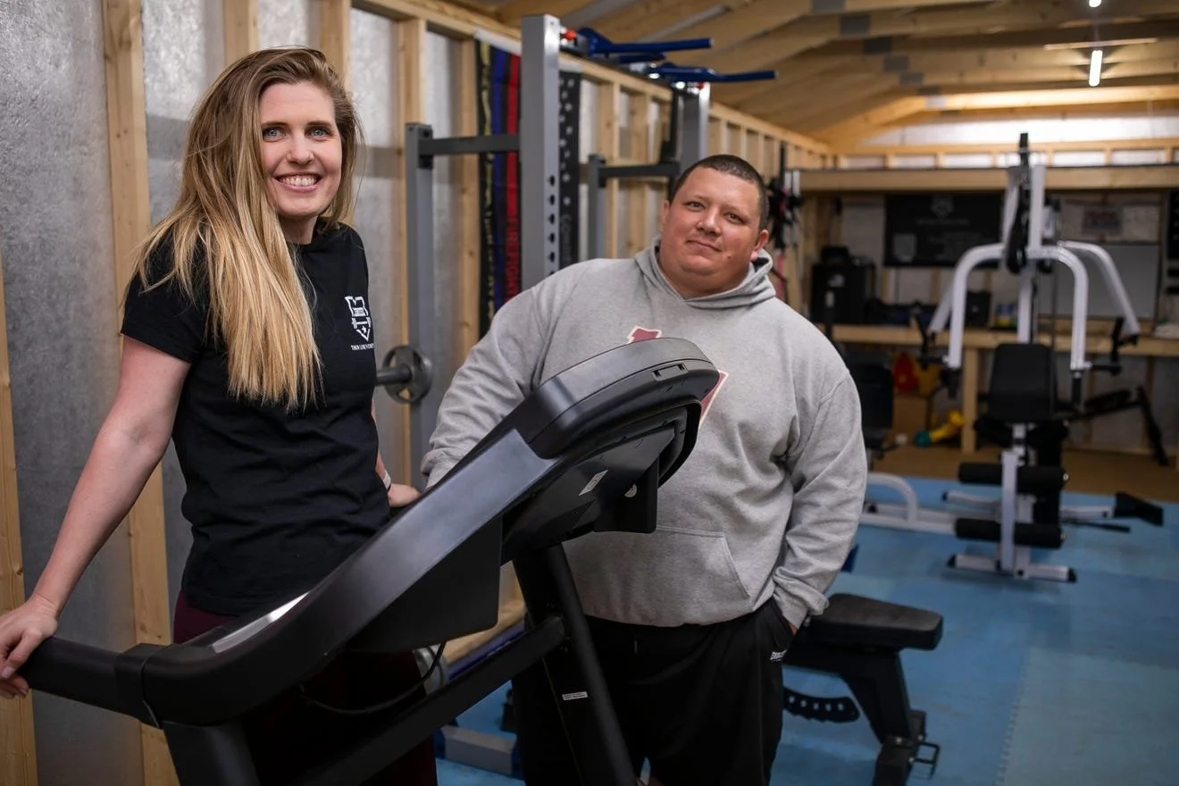 A woman and man standing in a gym, smiling at the camera. The woman is leaning on a treadmill, and the man is standing next to her in a gray hoodie. The gym has workout equipment and wooden walls.