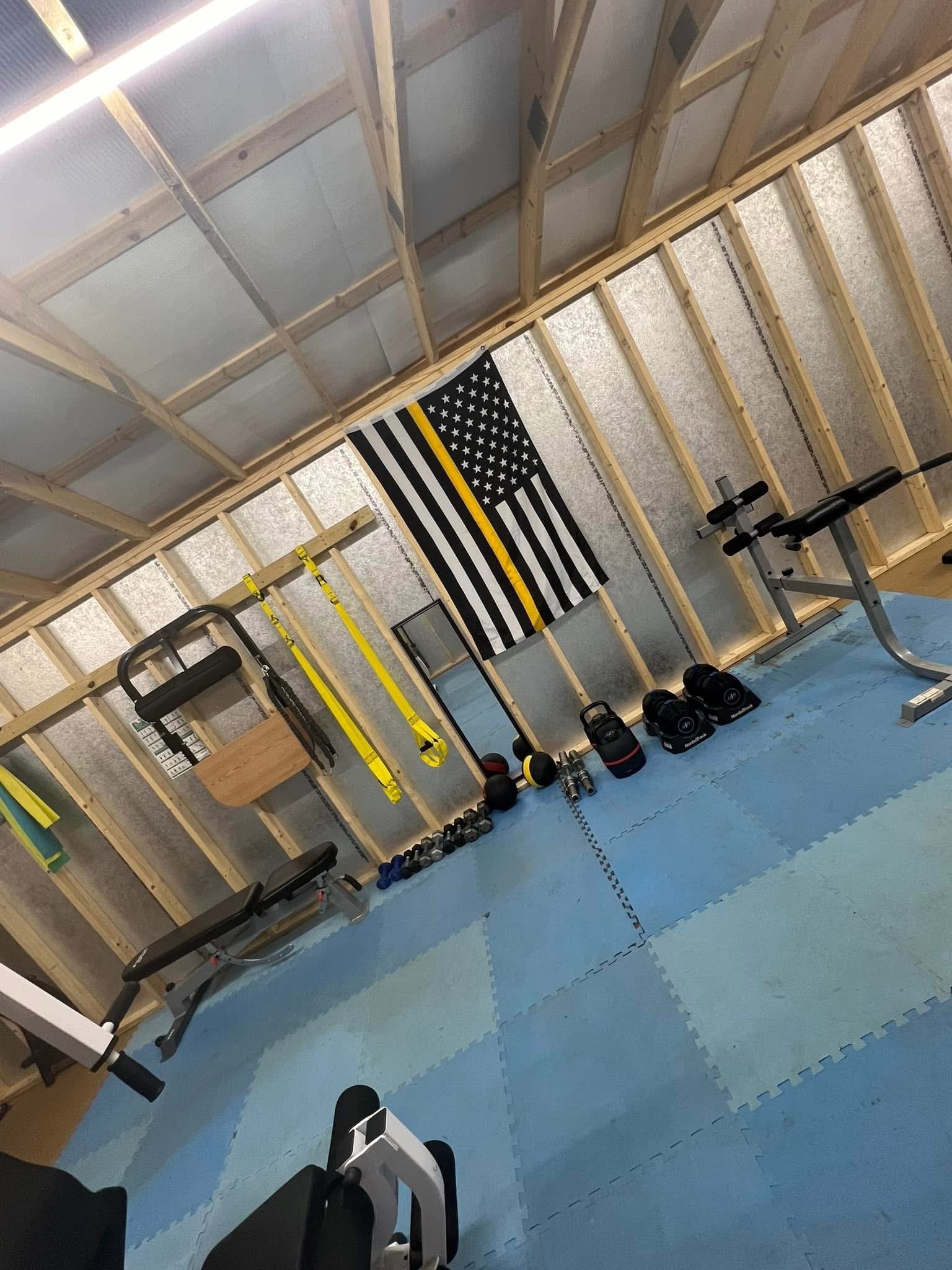 Home gym with exposed wooden framing, black and white American flag with a thin yellow line, various workout equipment including kettlebells, dumbbells, a bench, and a rowing machine, on blue interlocking foam mats.