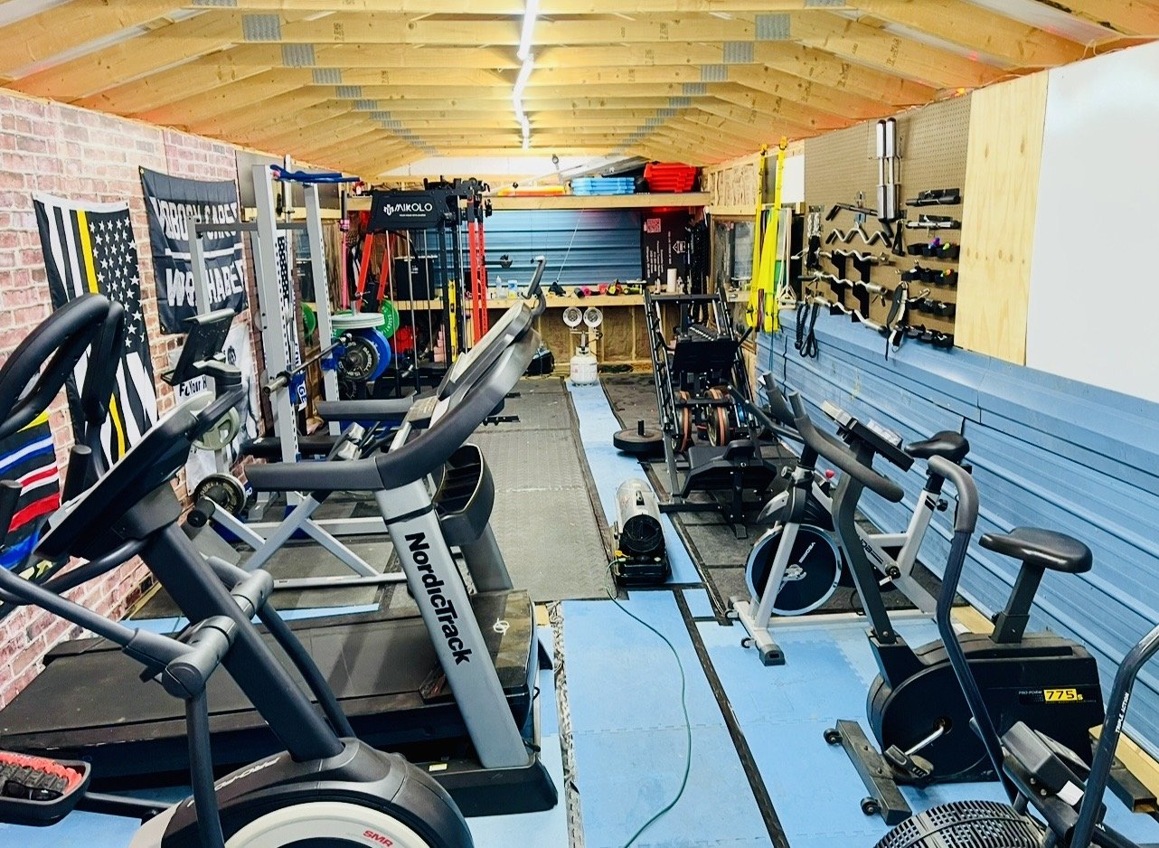 A well-organized home gym with exercise bikes, a treadmill, weight racks, and various fitness equipment against a brick wall and wooden walls with hooks for gym accessories.