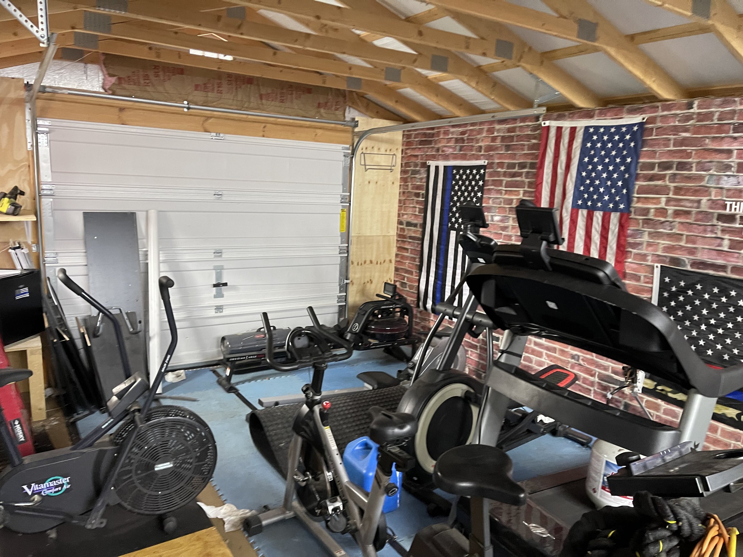 Home gym with exercise equipment in front of a brick wall with American flags and a black and white American flag. Includes a treadmill, exercise bike, and elliptical trainer in a garage with a partially open white garage door.