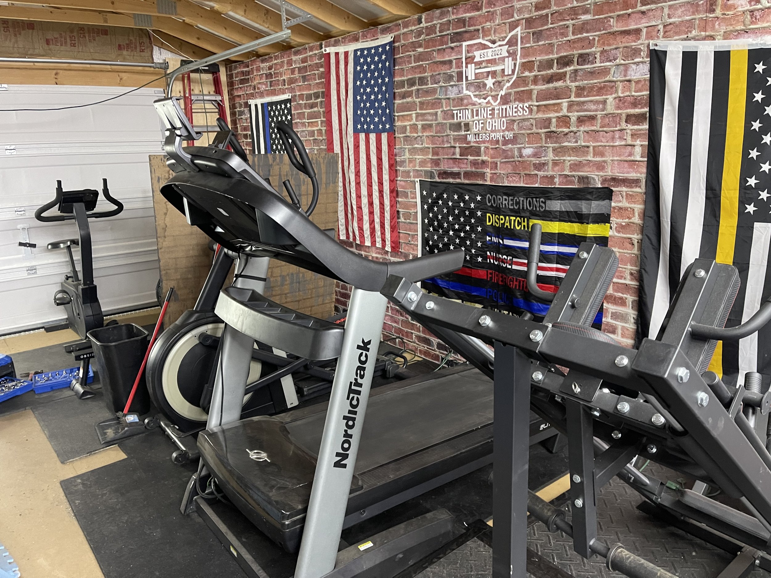 Indoor gym with a treadmill and an elliptical exercise machine, against a brick wall decorated with American flags and a correctional officer pride banner.